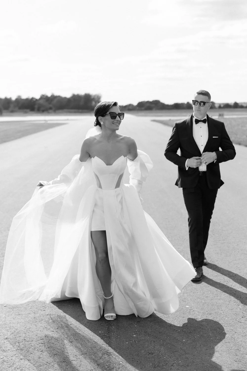A bride and groom walking on a runway outdoors, both wearing sunglasses and formal wedding attire, with the bride smiling and the groom holding a small item.