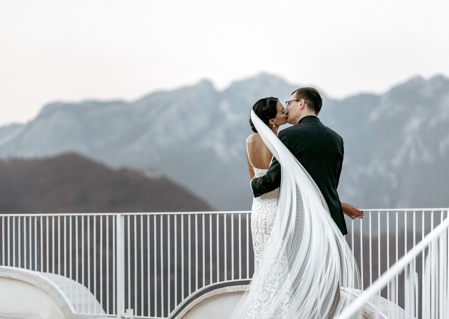 A newlywed couple sharing a kiss on a balcony with mountains in the background. The bride wears a white lace wedding gown and veil, while the groom is dressed in a black suit.