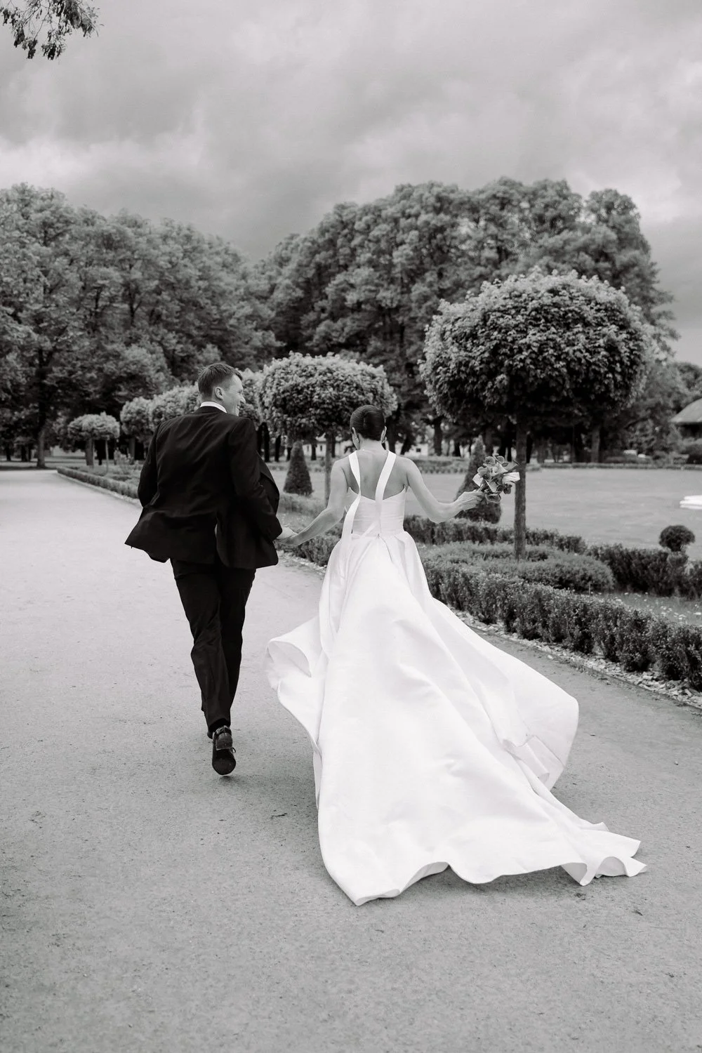 Black and white photo of a bride and groom walking away together in a park, with the bride holding a bouquet of flowers, trees and cloudy sky in the background.
