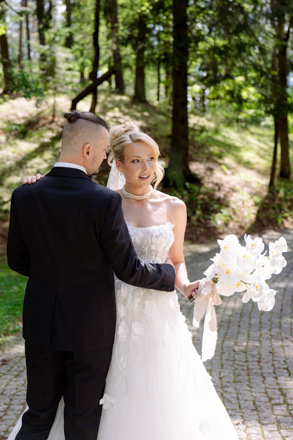 A bride and groom stand close together in a wooded outdoor setting during their wedding. The bride wears a white strapless wedding gown and pearl necklace, holding a bouquet of white orchids. The groom is dressed in a black suit and has his hair tied