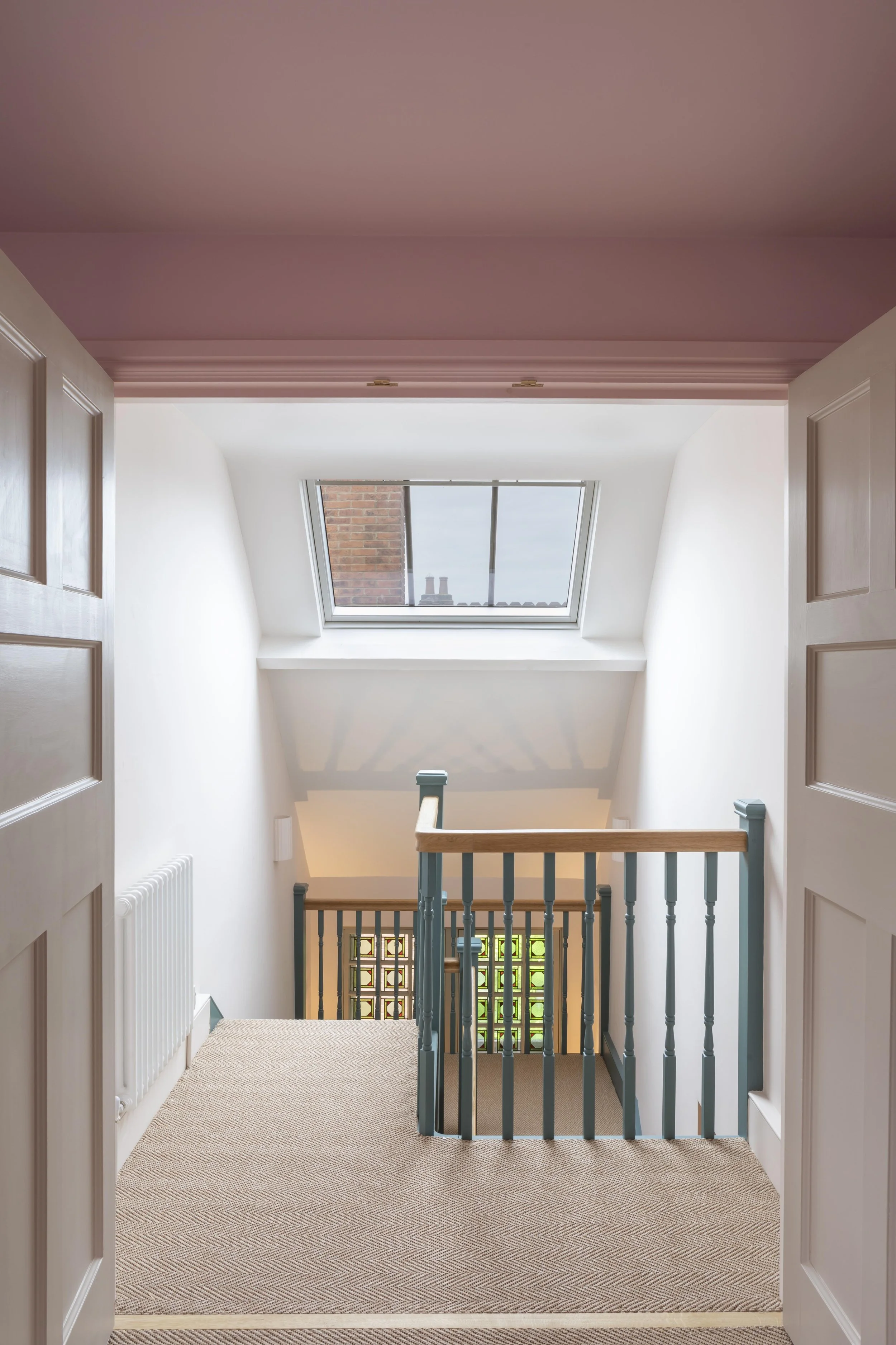 Interior view of a stairway landing serving a second floor Loft conversion, with a skylight window above the stair, white walls, a small radiator on the left, and blue railings with wooden handrails. The carpeted floor is beige with a subtle pattern.