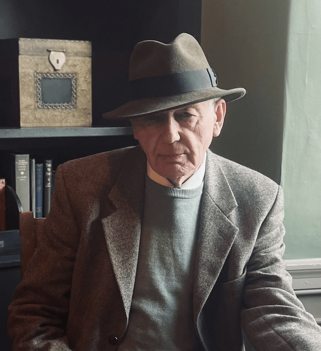 Nigel, the author, wearing a brown fedora hat, a grey sweater, and a brown tweed blazer, sitting in a room with books and a decorative box in the background.