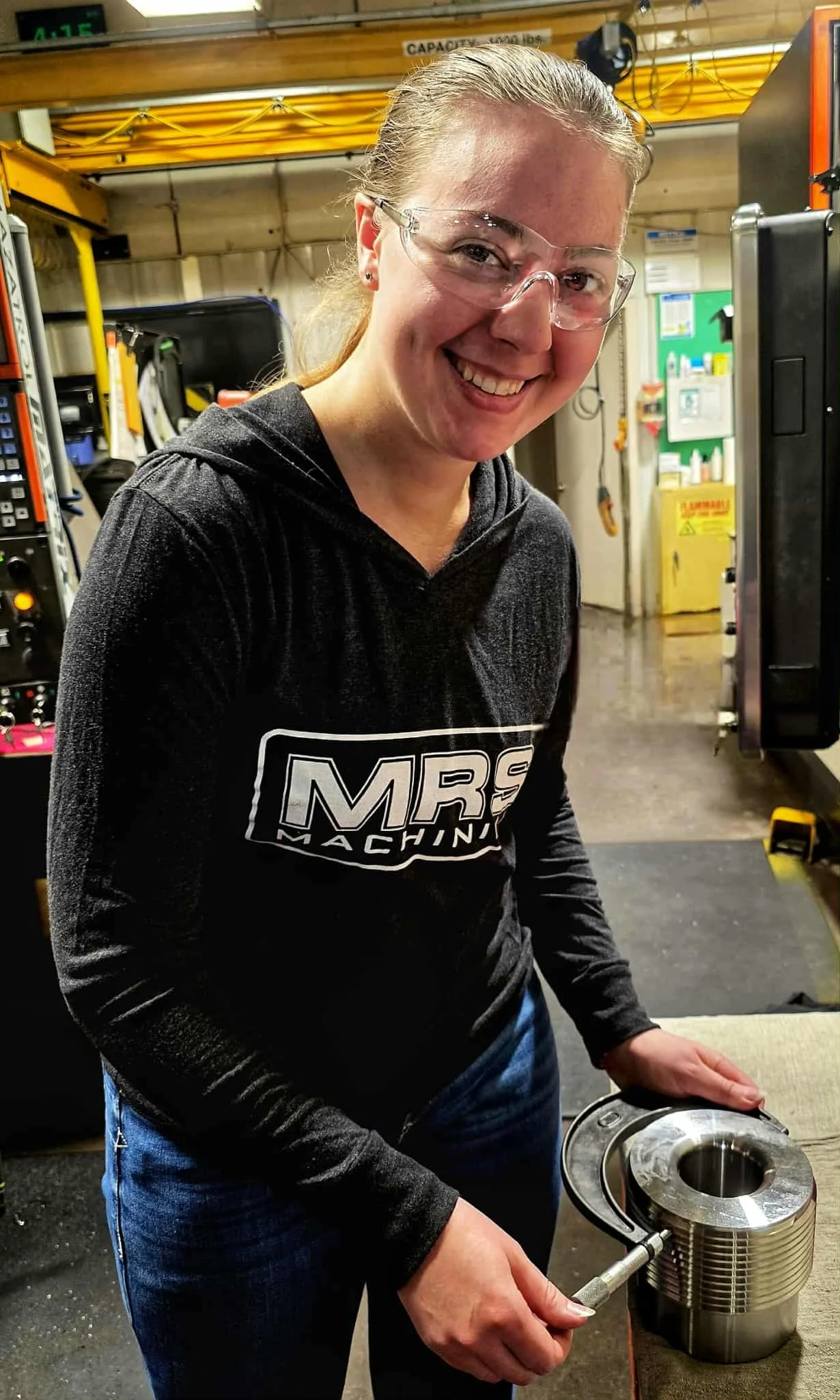 A woman with safety glasses and a smile, working in a workshop, holding a metal component with a tool, wearing a black hoodie with 'MRS MACHINING' printed on it, surrounded by industrial equipment.