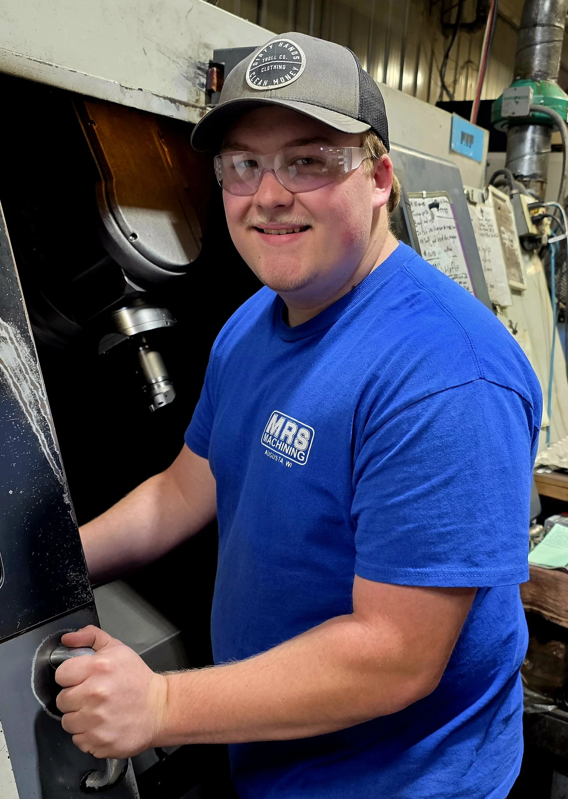A young man wearing safety glasses, a baseball cap, and a blue t-shirt with the MRS Machining logo, working at a CNC machine in a workshop.