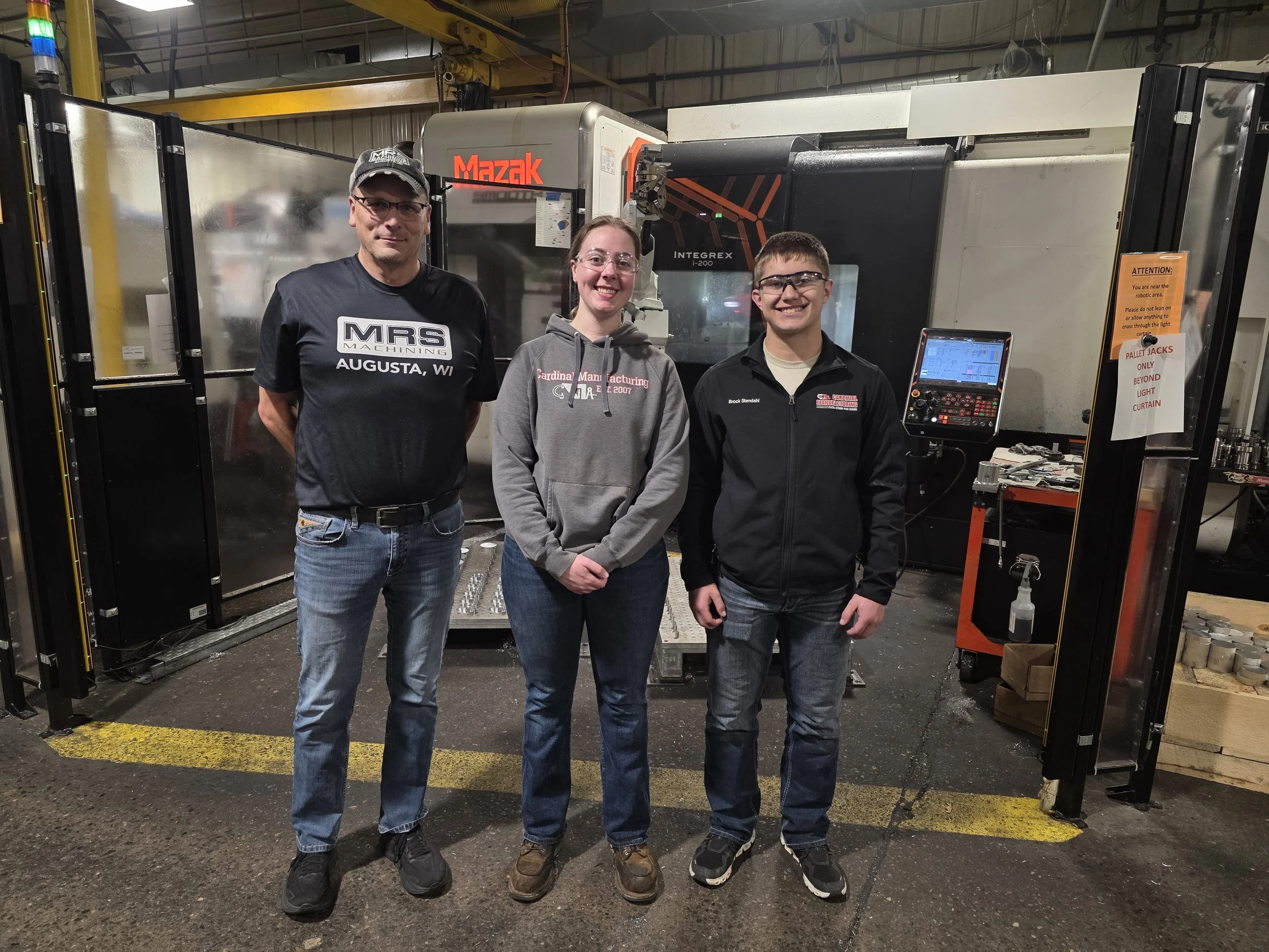 Three people standing in an industrial workshop in front of a large CNC machine. The person on the left is wearing a black t-shirt, jeans, and a cap. The person in the middle is wearing a grey hoodie and jeans, and the person on the right is wearing 