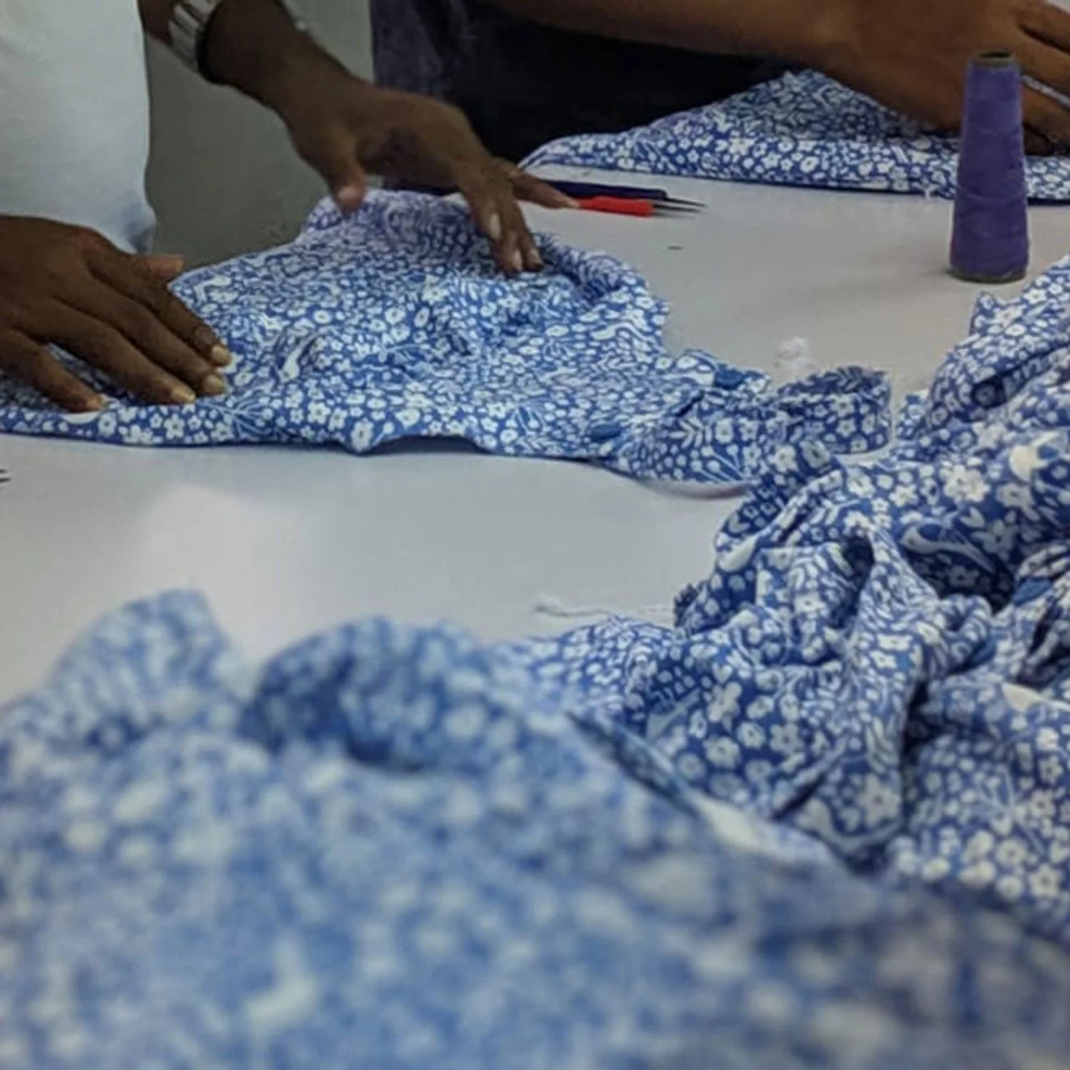 Blue floral girls dungaree dress having buttons applied by hand in factory there is a spool of purple blue thread to one side