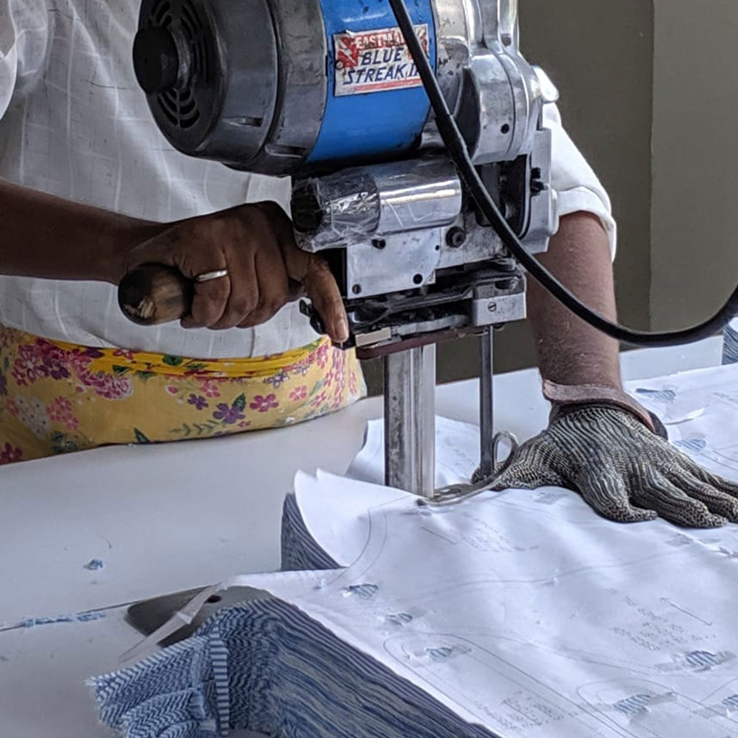 Kidswear pattern pieces being cut by machine out of blue striped fabric the person operating the machine is wearing a white shirt and yellow floral pinny