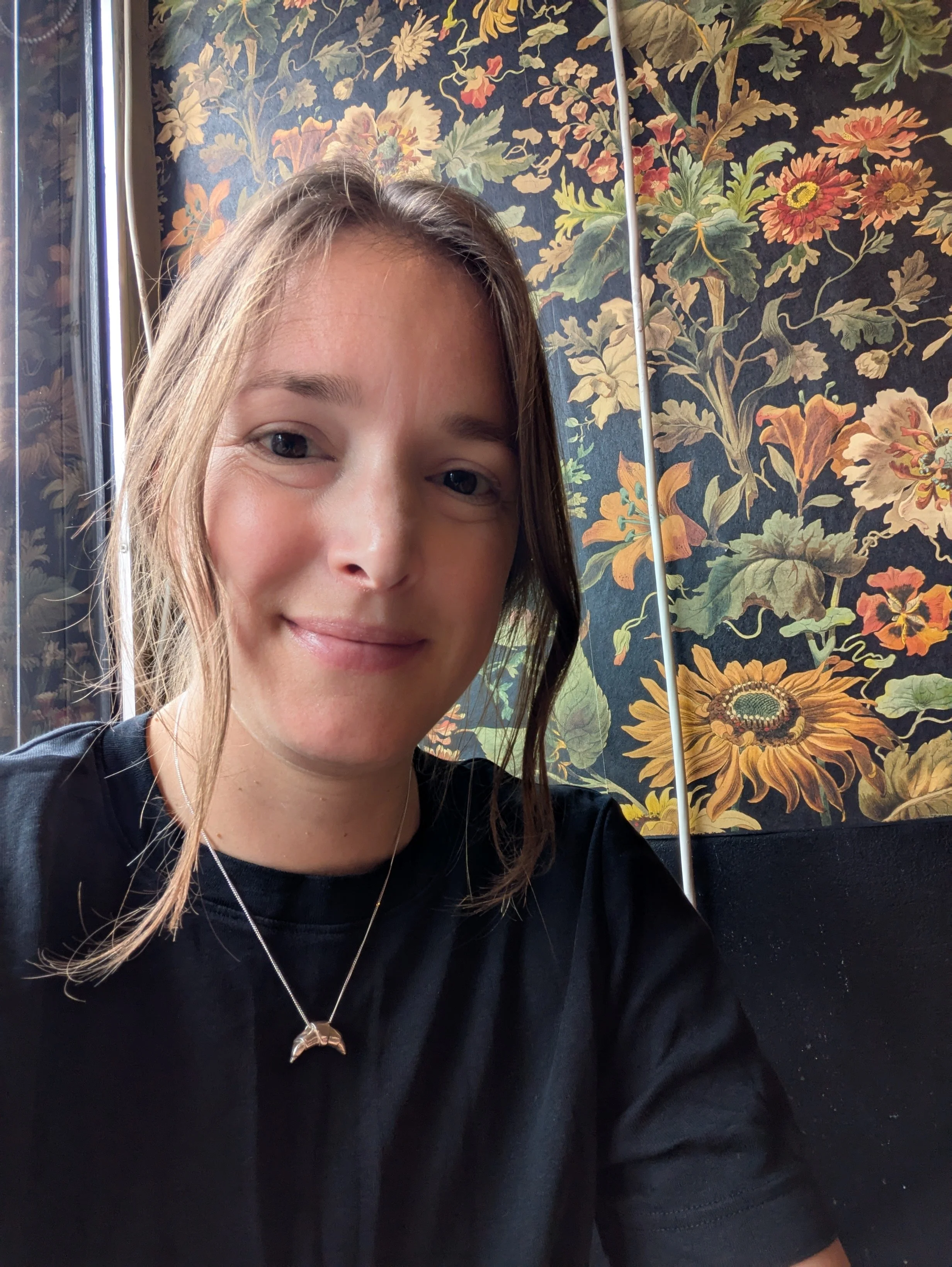Childrenswear designer Sherry Pritchet, smiling at the camera, wearing a black shirt and a necklace with a croissant pendant. Behind her is a window with floral-patterned wallpaper featuring sunflowers, roses, and other flowers.