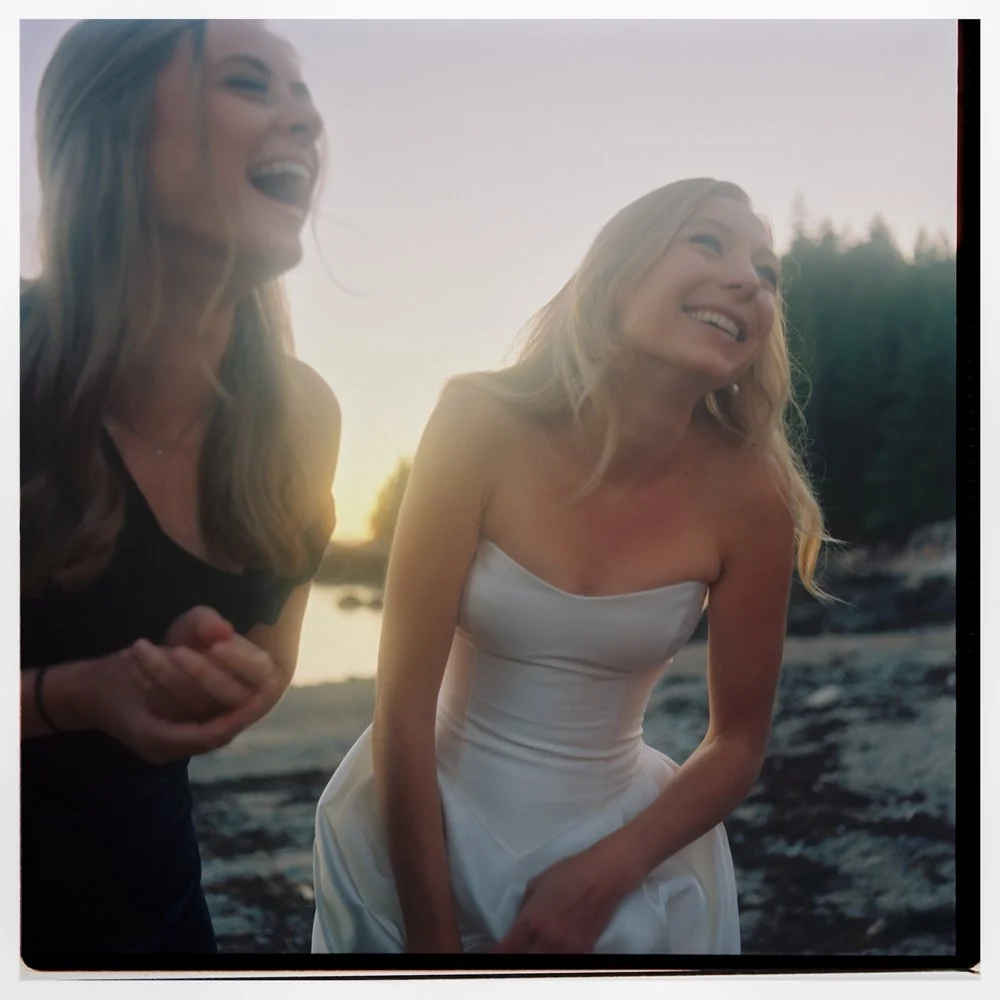 Two women laughing outdoors near a river at sunset, one in a black top, the other in a white strapless dress. Film wedding photographer in Maine. 