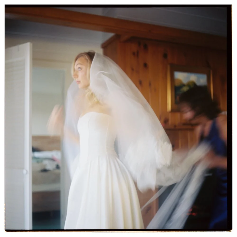 A bride in a white wedding dress adjusting her veil inside a room with wooden walls, with a woman in blue helping her. Film wedding photographer in Maine. 
