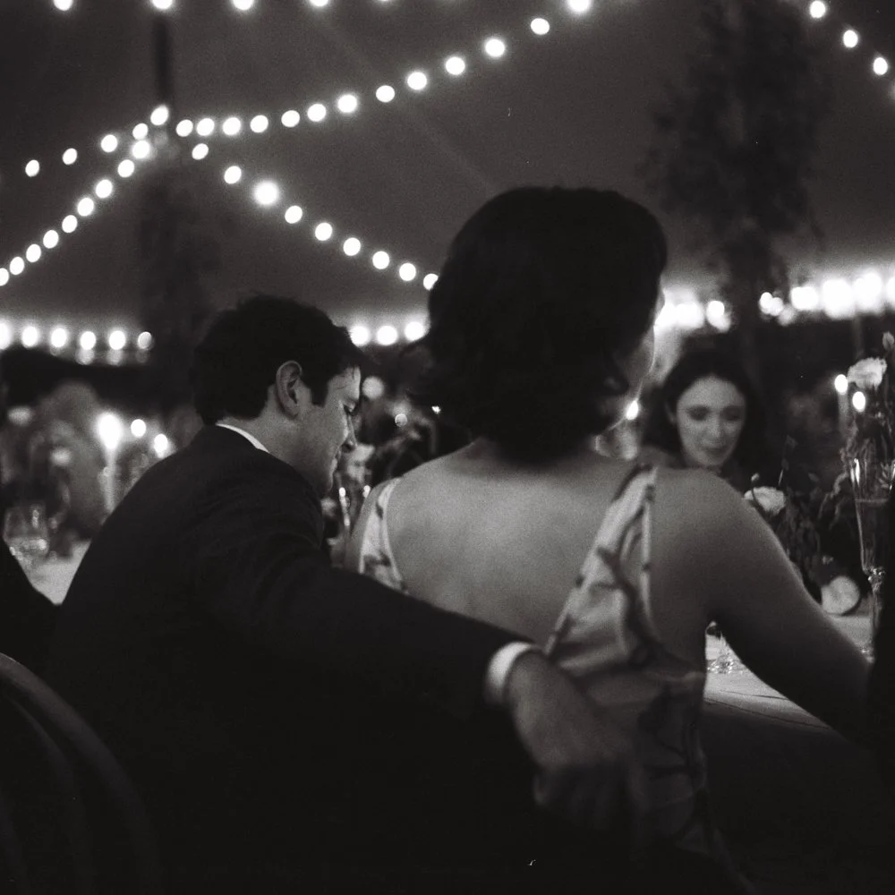 People sitting at a nighttime outdoor celebration with string lights overhead. Non -traditional film wedding photographer in Maine. 
