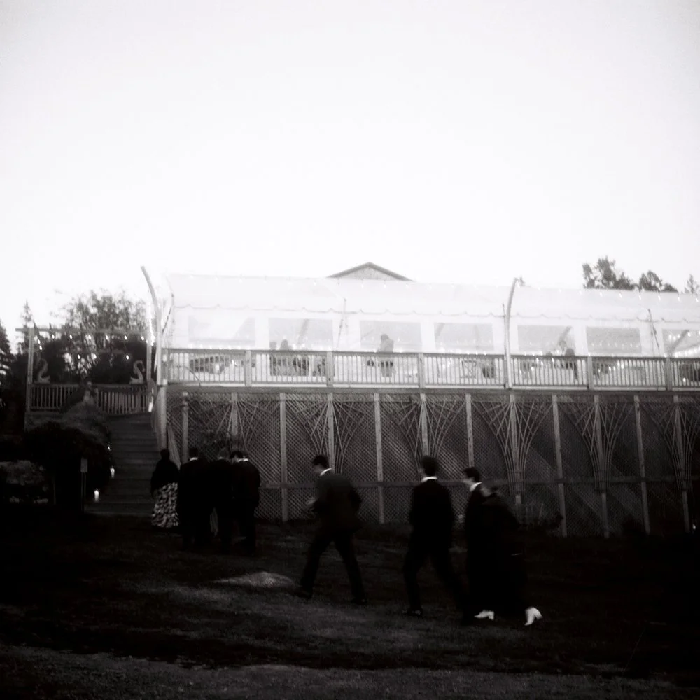 A group of people walking down a grassy hill past a wooden fence, with a large elevated structure with a white canopy and people inside on the upper level, visible in the background. Film wedding photographer in Maine. 