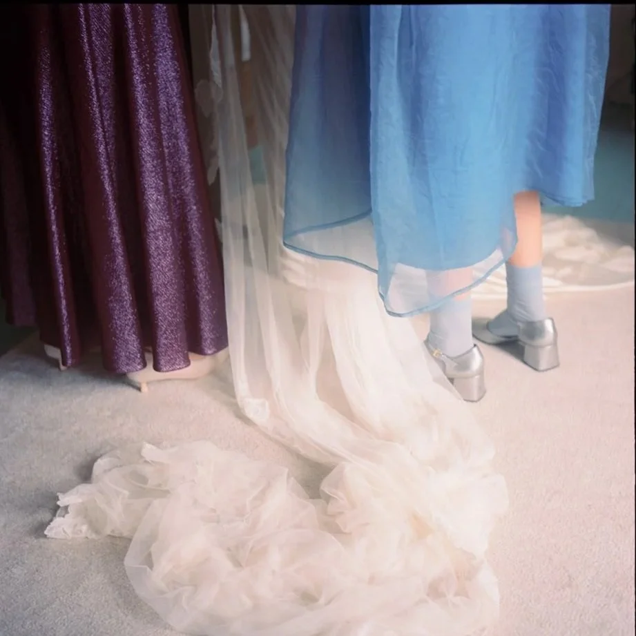 A person wearing a blue dress and silver platform shoes with socks, standing amidst dresses and a train of a wedding gown on the floor.