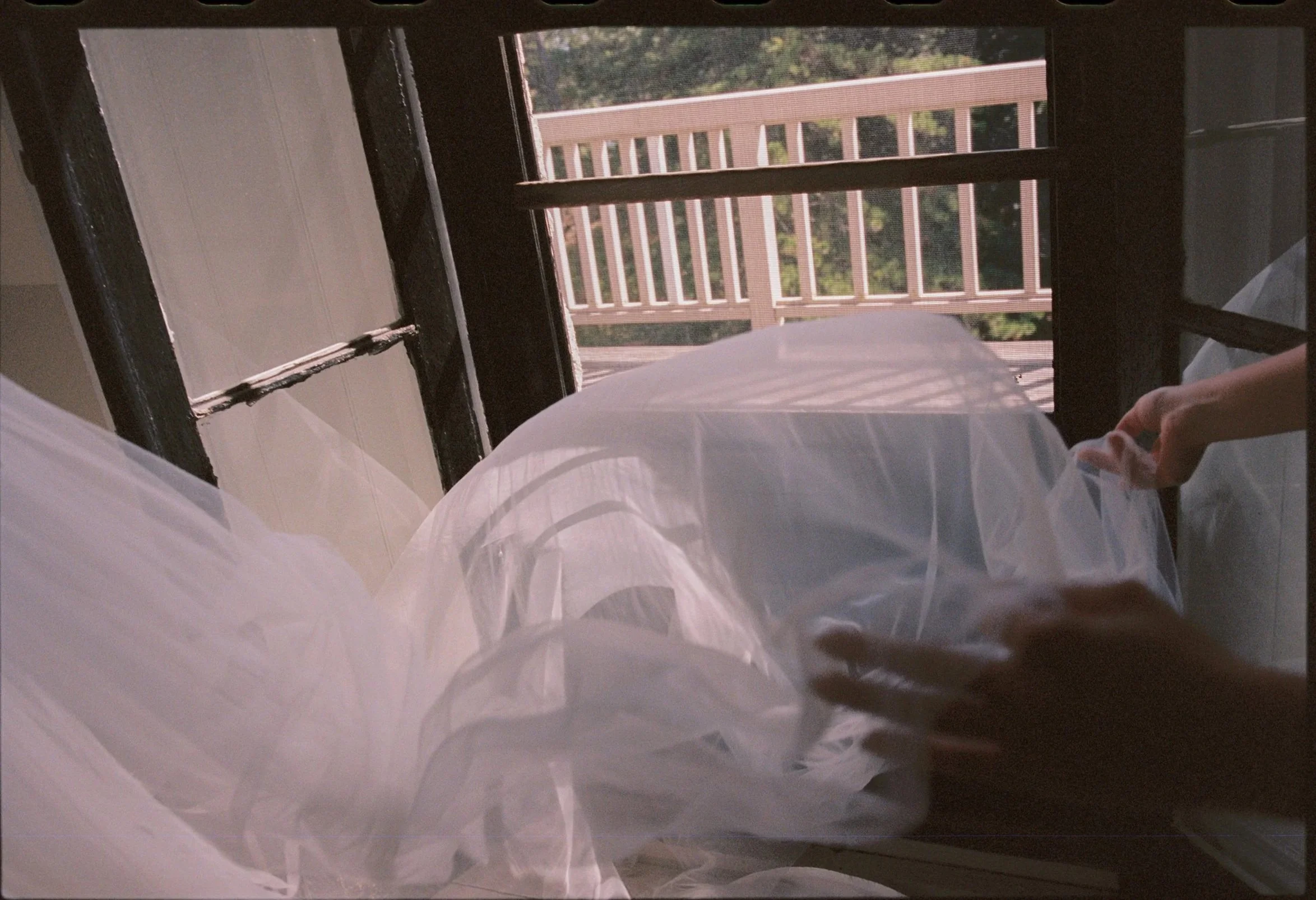 A person is holding a white fabric or plastic sheet near a window with sunlight streaming through, revealing an outdoor balcony with a pink railing.