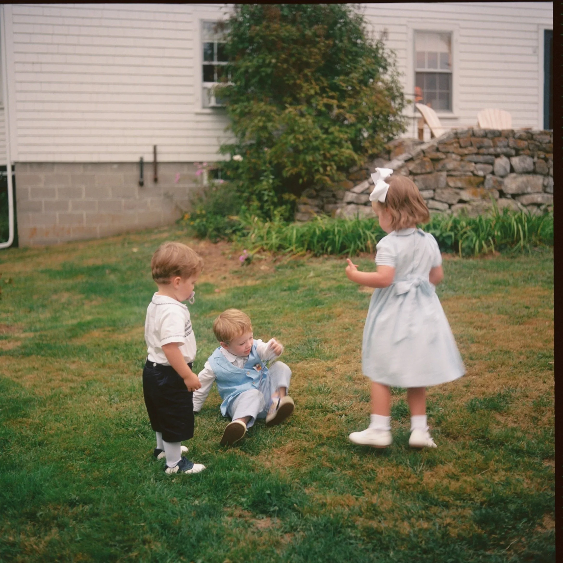Three young children playing on a lawn in front of a house, with two girls and one boy, during daytime. Film wedding photographer in Maine. 