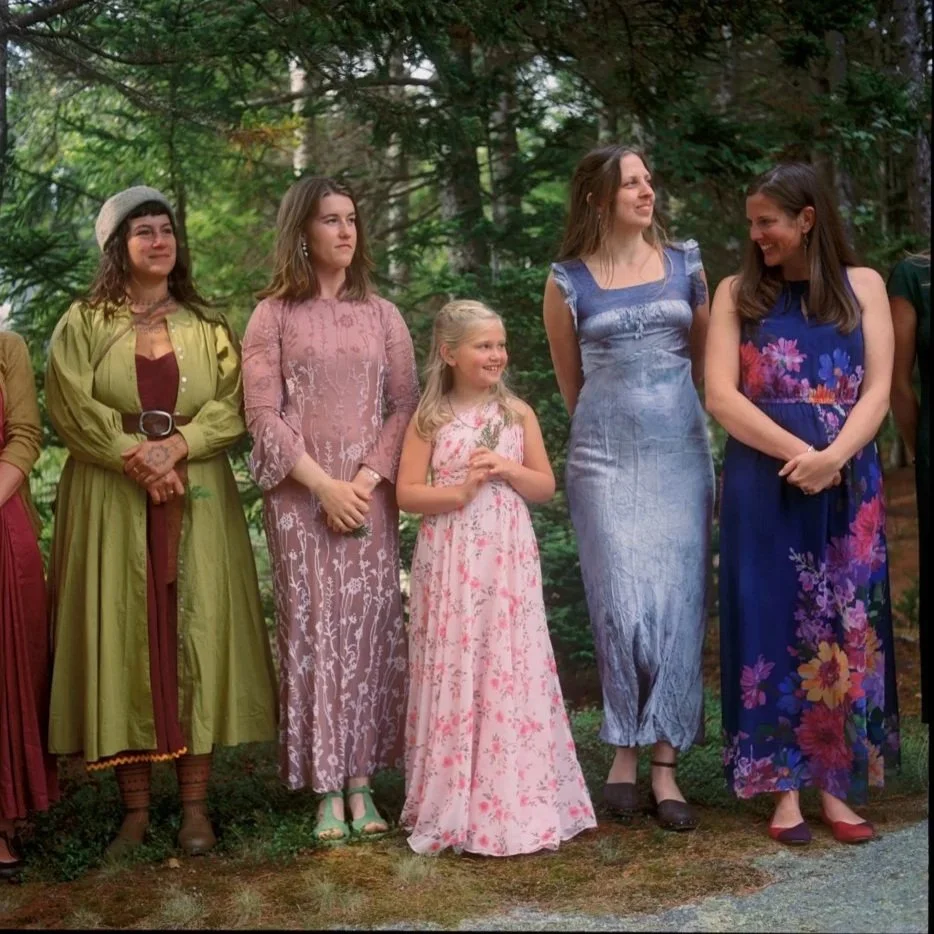 Group of women and a girl standing outdoors in a wooded area, dressed in colorful dresses, smiling and looking in different directions.