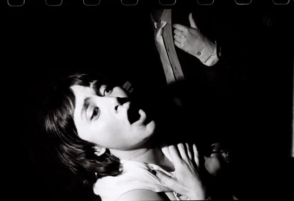 A black and white photo of a woman with short hair laying down, with her mouth open and hand on her chest. A person in a suit is partially visible next to her, adjusting their tie.