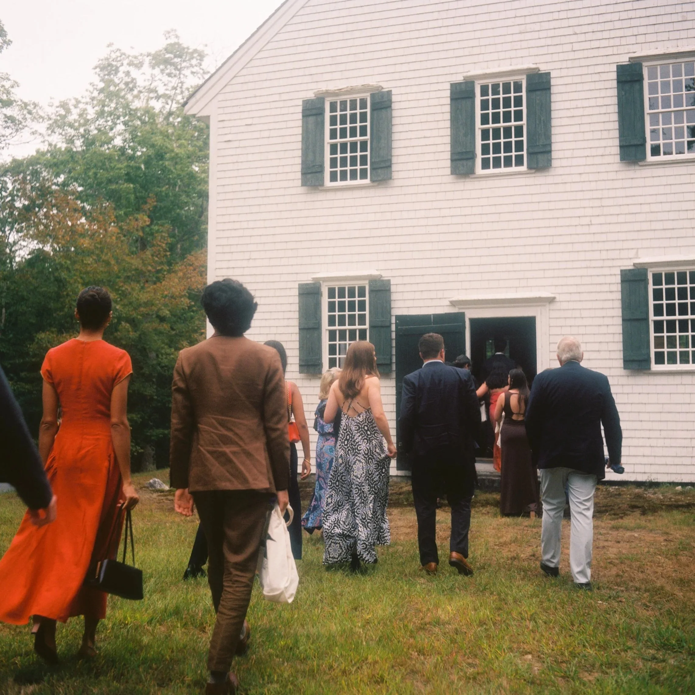 People dressed in formal attire are entering a large white colonial-style house with blue shutters for an event, with trees in the background and a grassy lawn in the foreground. Film wedding photographer in Maine. 