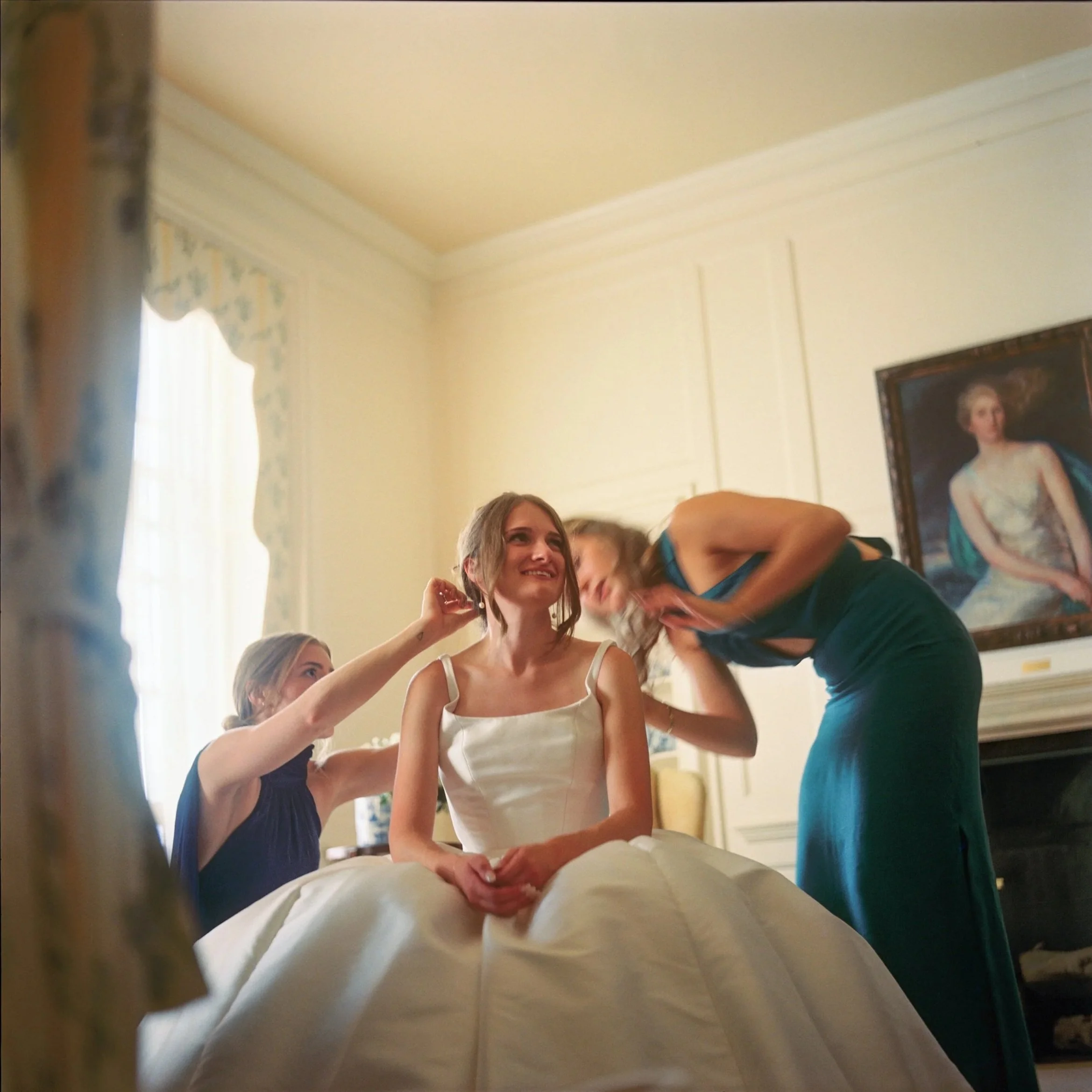 A bride sitting in a room with two women helping her prepare, likely for her wedding, as one woman adjusts her earring and the other touches her hair. The bride wears a satin wedding gown, and the room has elegant decor with a painting of a woman in 