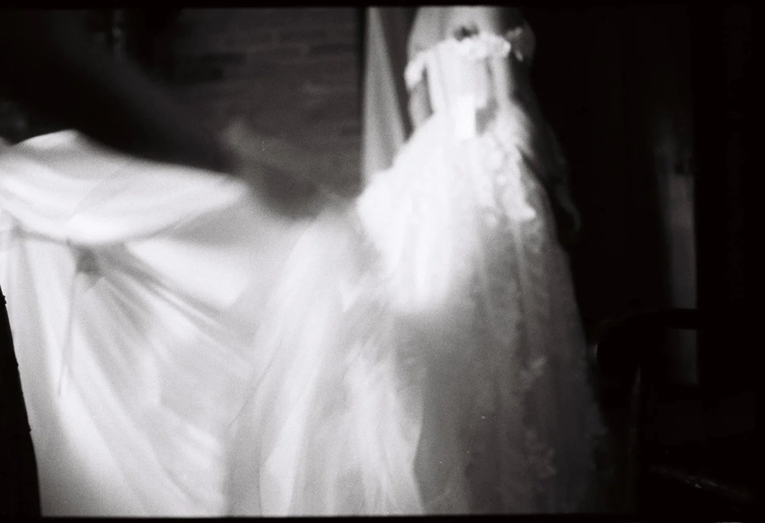 A blurred black and white photo of a wedding dress hanging, with some fabric flowing to the side. Film wedding photographer in Maine. 