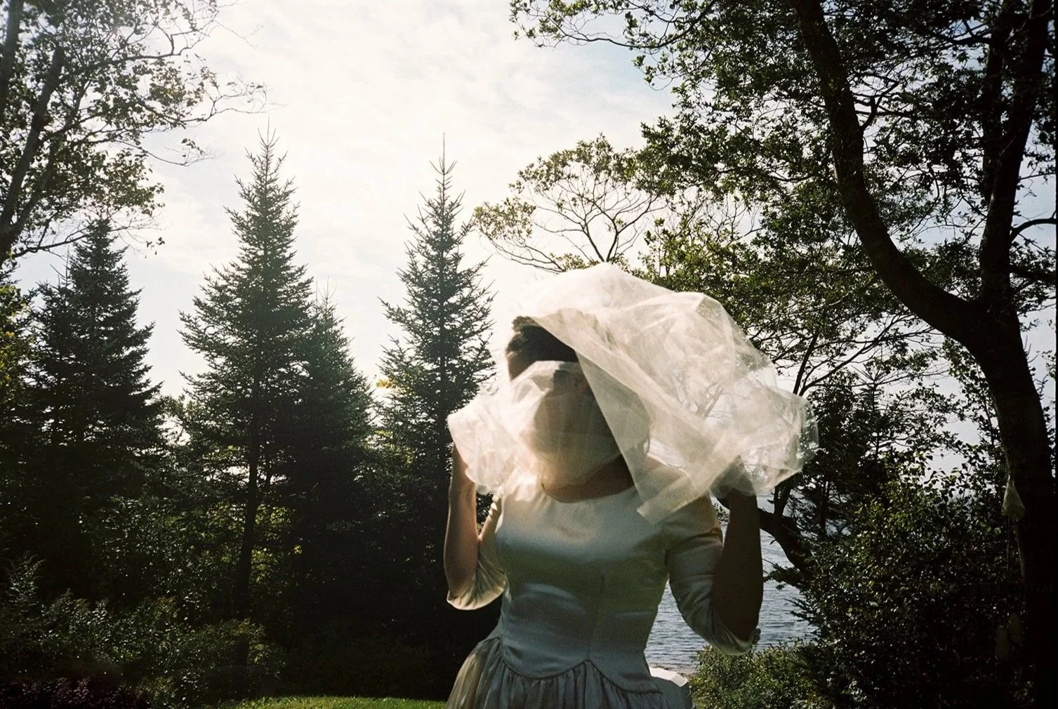 A woman in a vintage white dress and large bow hat standing outdoors in a wooded area with trees and water in the background, backlit by sunlight.