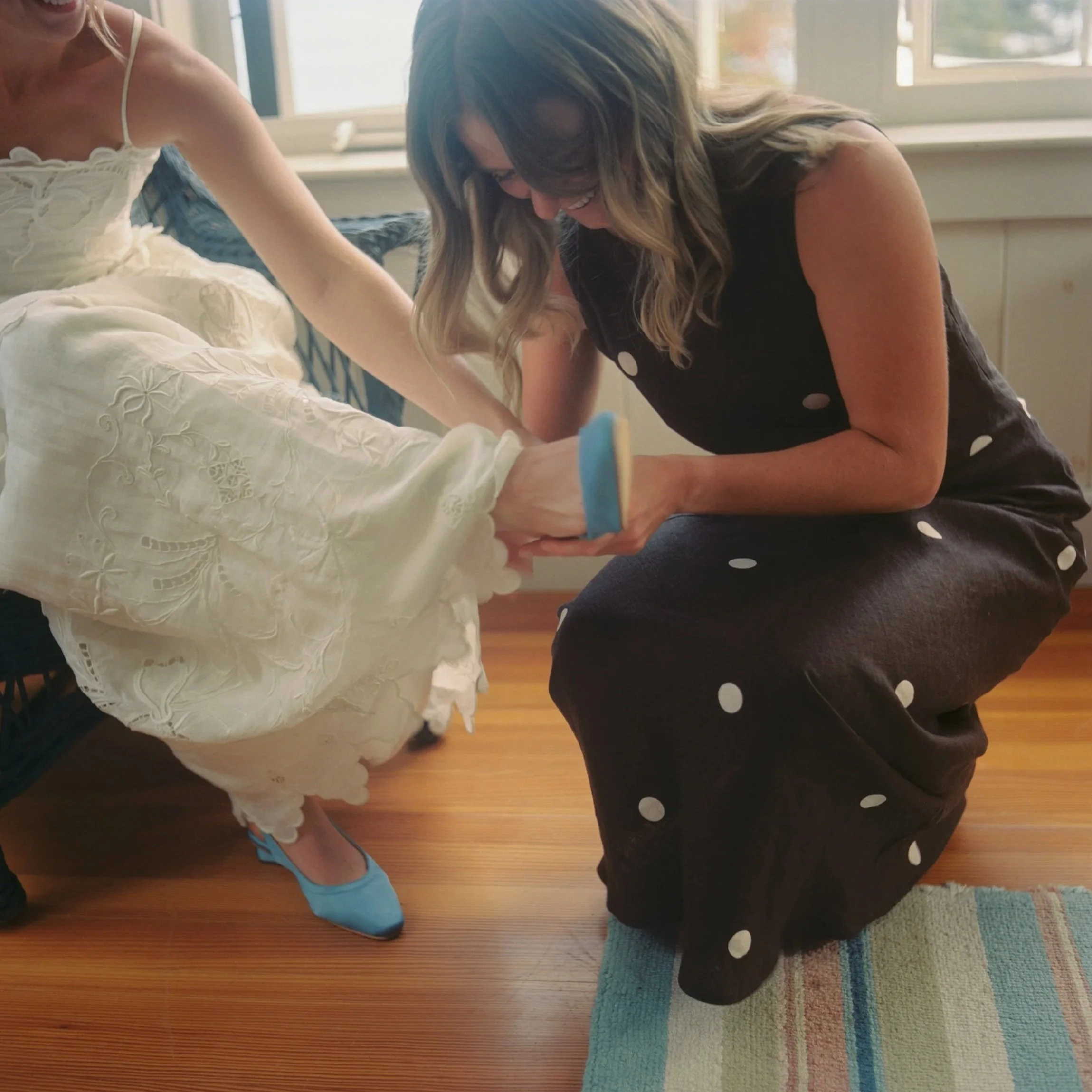 Two women, one in a white embroidered dress and the other in a black polka dot dress, are laughing and holding hands. They are inside near a window with sunlight coming in, on a hardwood floor with a colorful striped rug.