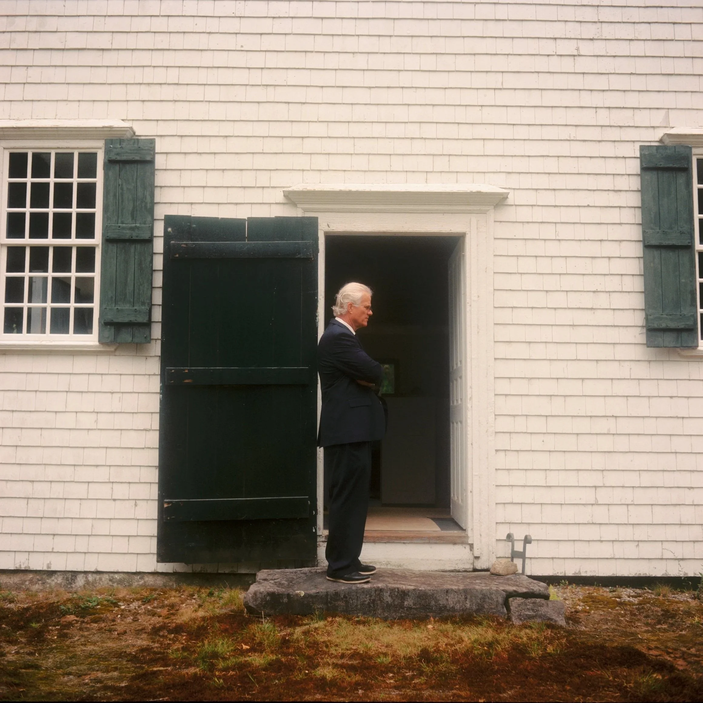 A man in a black suit standing with arms crossed on a small porch in front of a white house with green shutters.