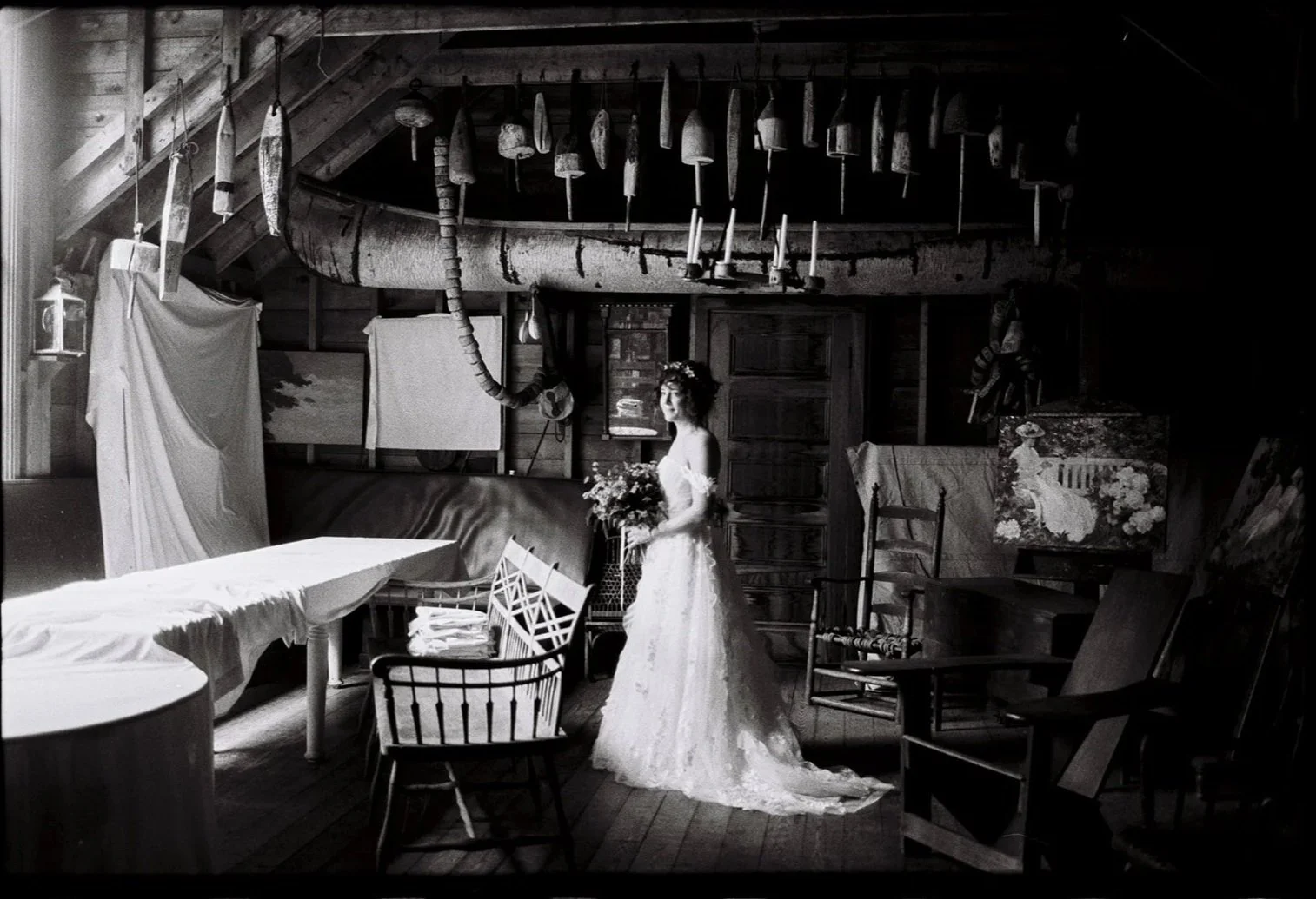 Black and white photo of a woman in a wedding dress holding a bouquet in a rustic room with wooden walls, various hanging tools, artwork, and furniture. Film wedding photographer in Maine. 