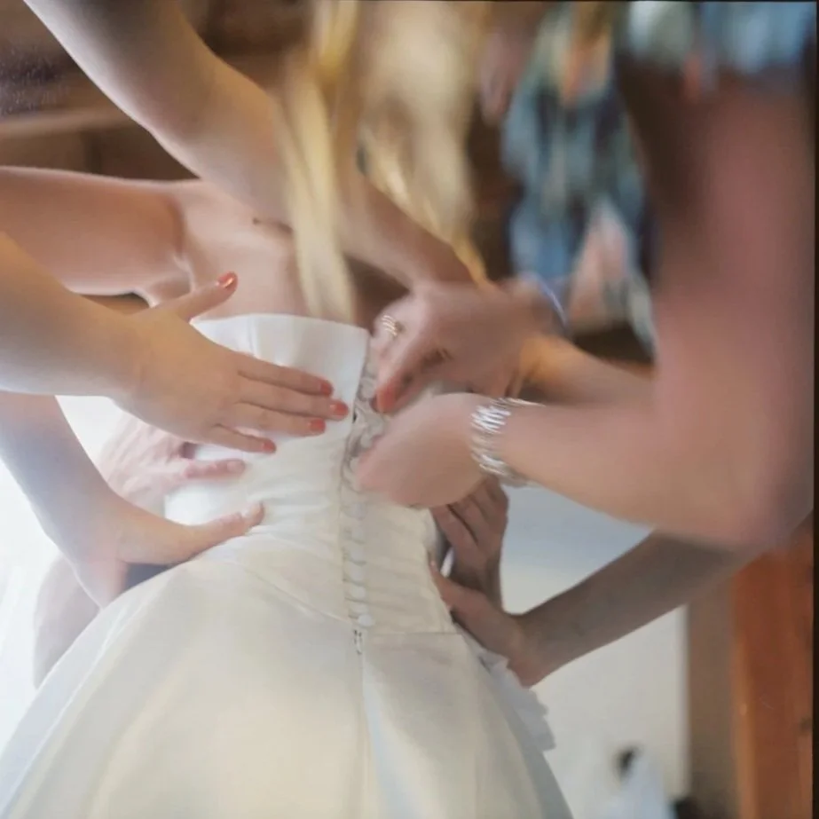Close-up of a bride in a wedding dress being assisted by several women to zip up the back of her gown. Film wedding photographer in Maine. 