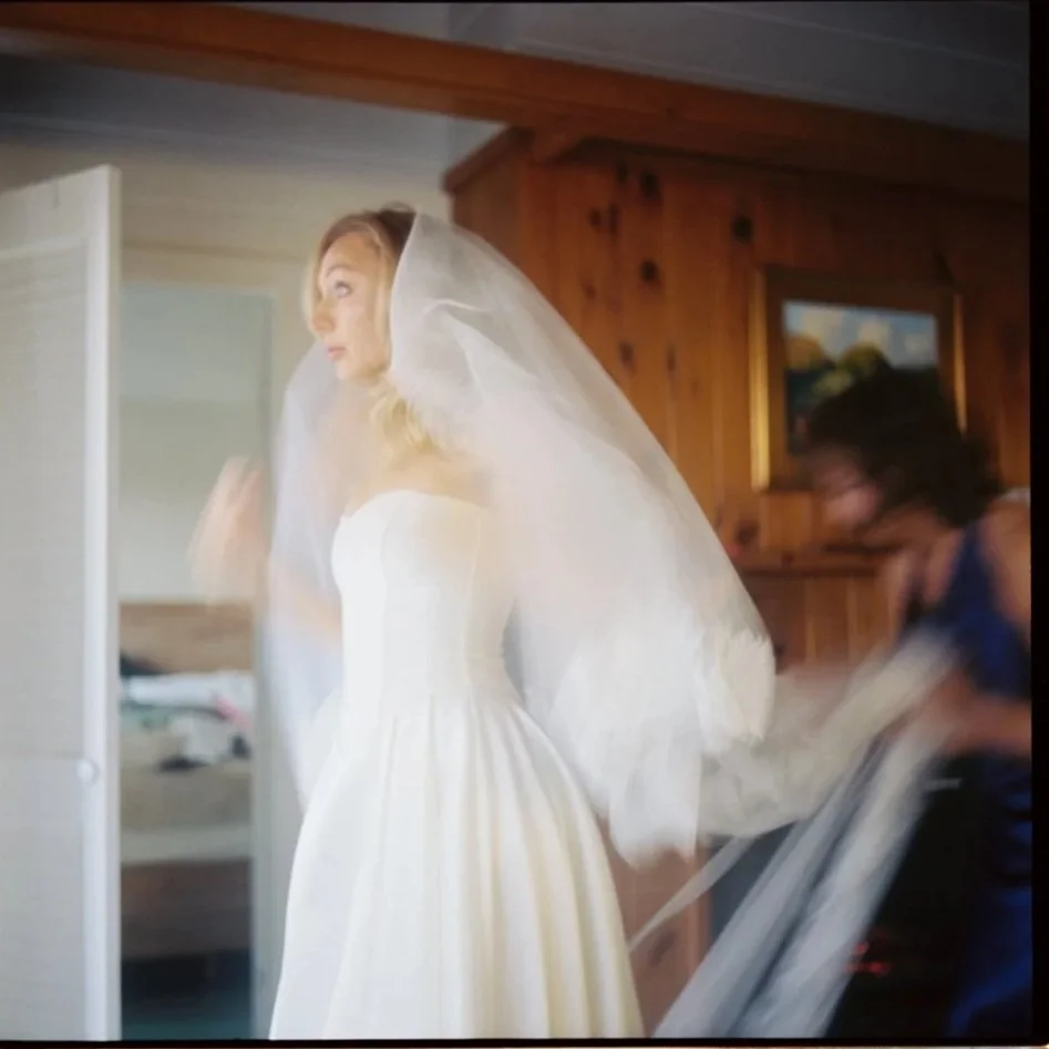 A bride in a white wedding dress adjusting her veil inside a room with wooden walls, with a woman in blue helping her. Film wedding photographer in Maine. 