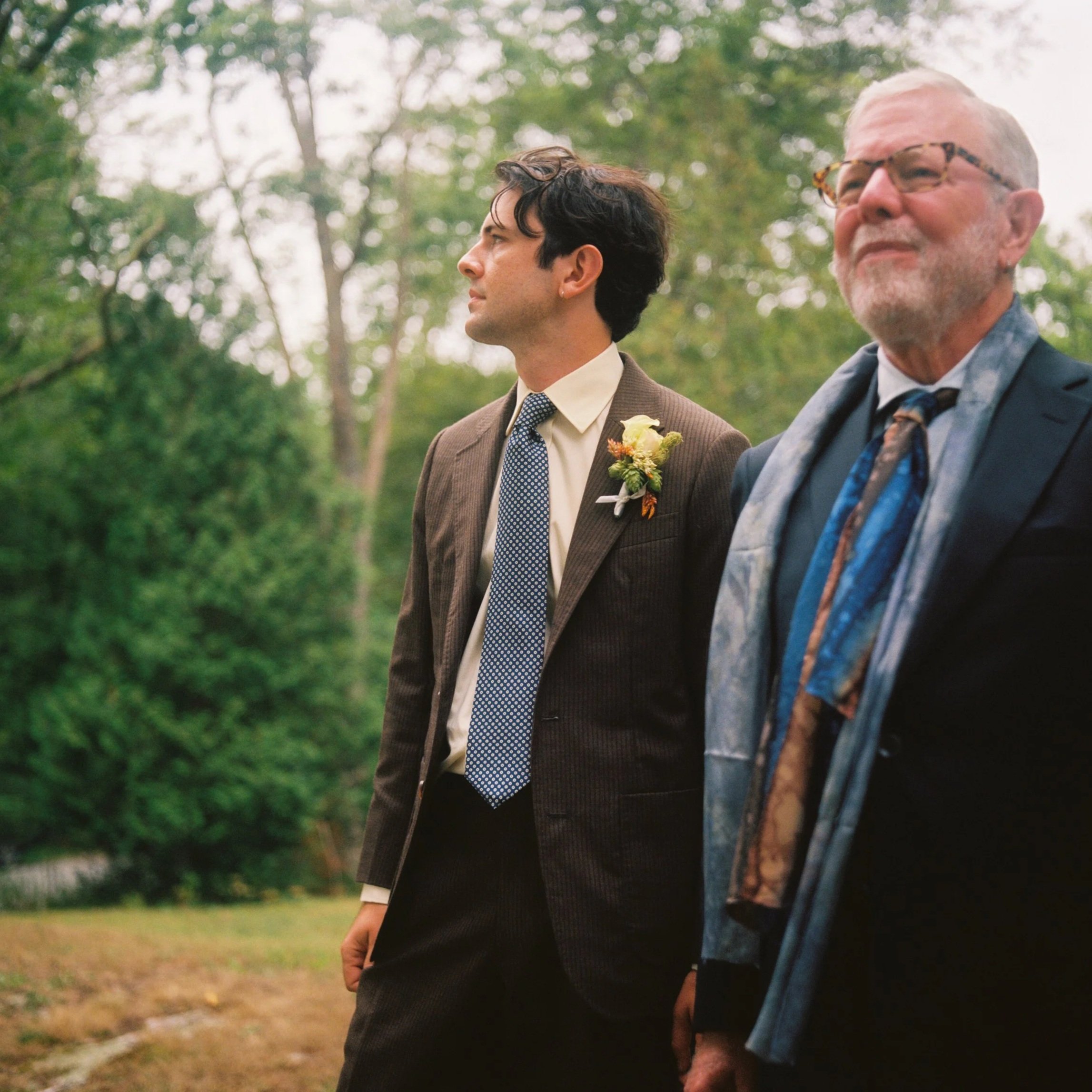 A young man and an older man standing outdoors in a park-like setting, both dressed in suits. The young man wears a brown suit with a white shirt, blue patterned tie, and a boutonniere. The older man wears glasses, a dark suit with a patterned tie, a