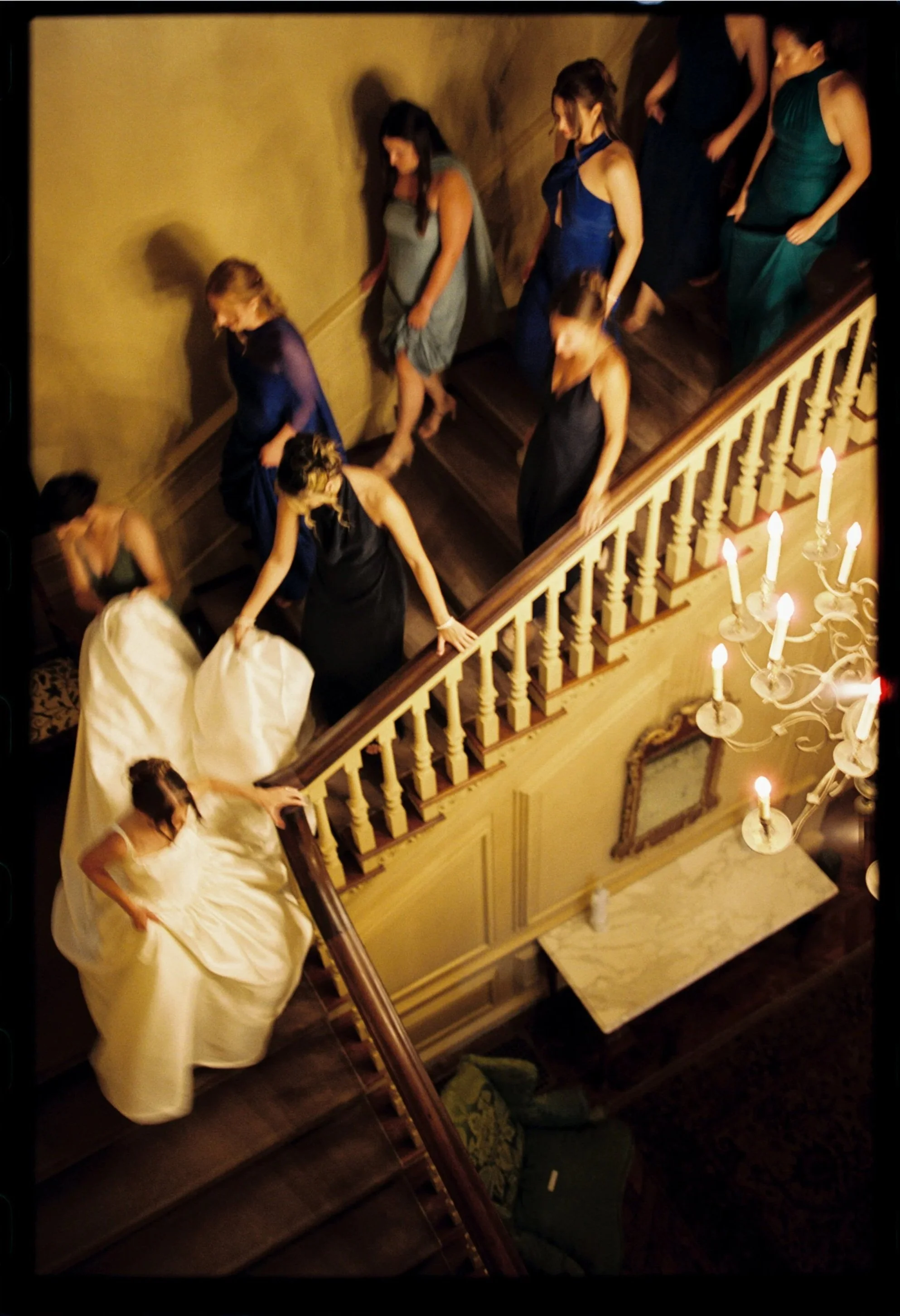 Women in formal evening gowns descending a grand staircase at a fancy event, with a chandelier hanging above. Film wedding photographer in Maine. 