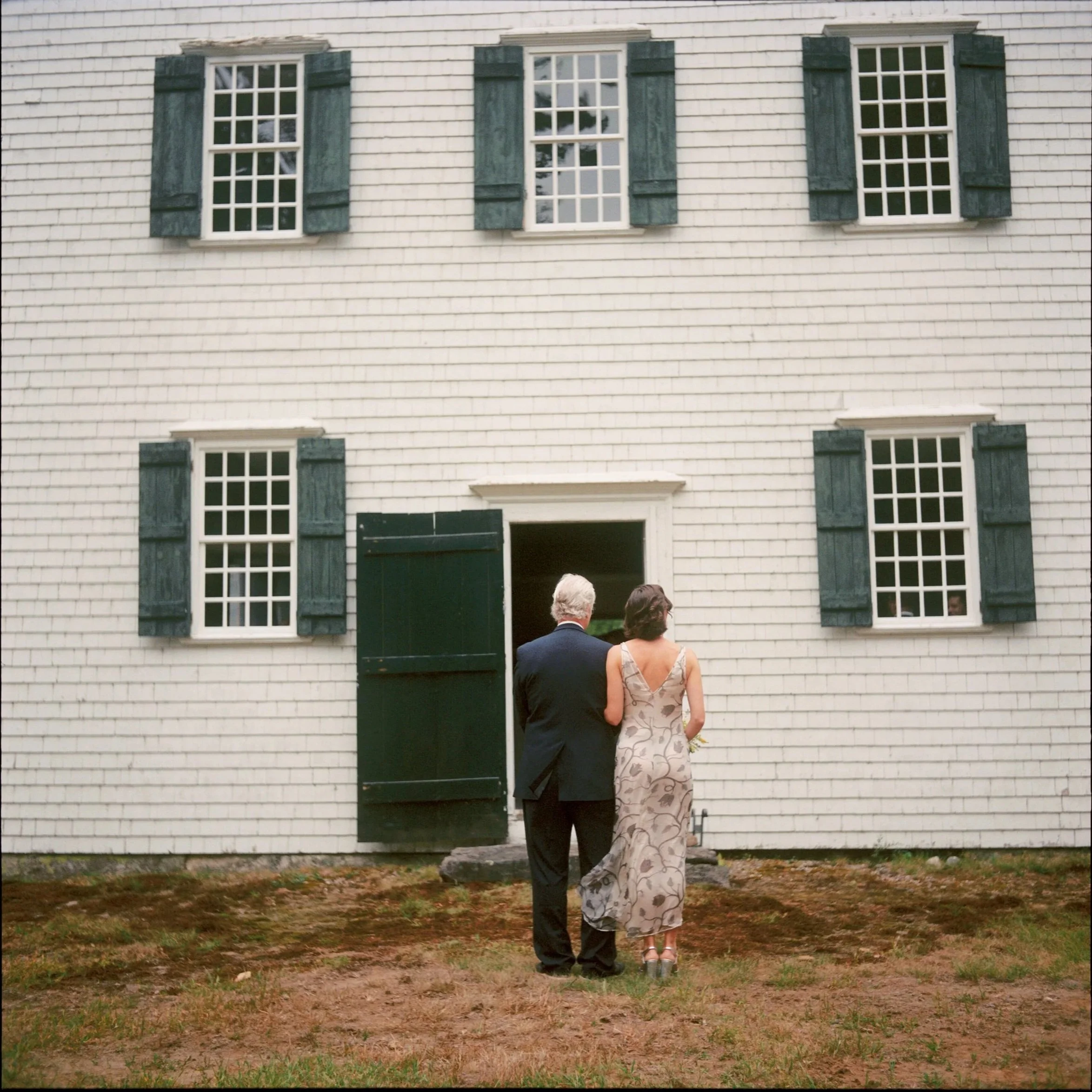 A couple, an older man in a suit and a woman in a floral dress, walking towards an open door in front of a white house with dark shutters. Film wedding photographer in Maine. 