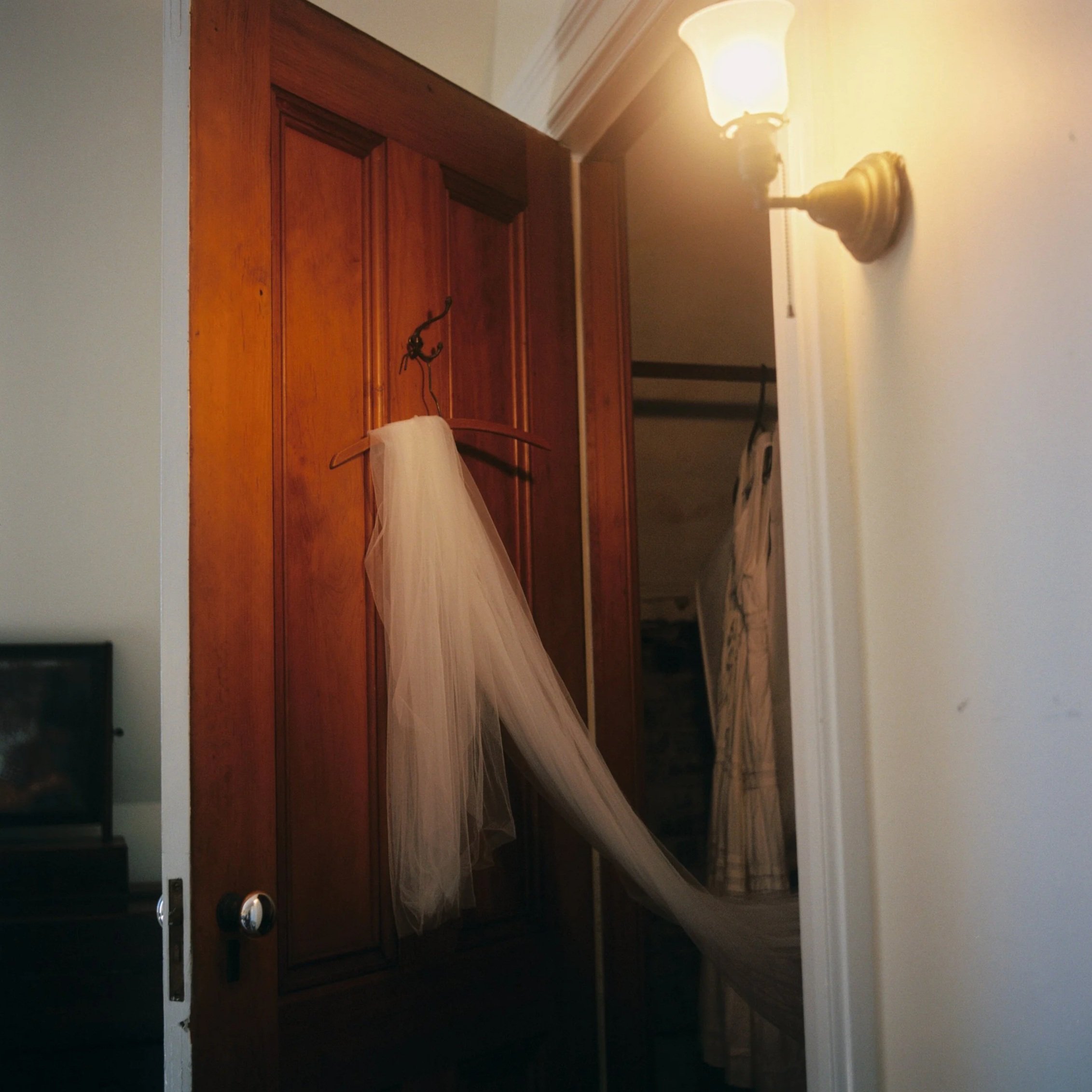 A cream-colored veil hanging on a wooden wardrobe door with a brass hook. A wall-mounted light fixture illuminates the scene. Part of a bedroom with a door leading to another room or closet in the background. Film wedding photographer in Maine. 