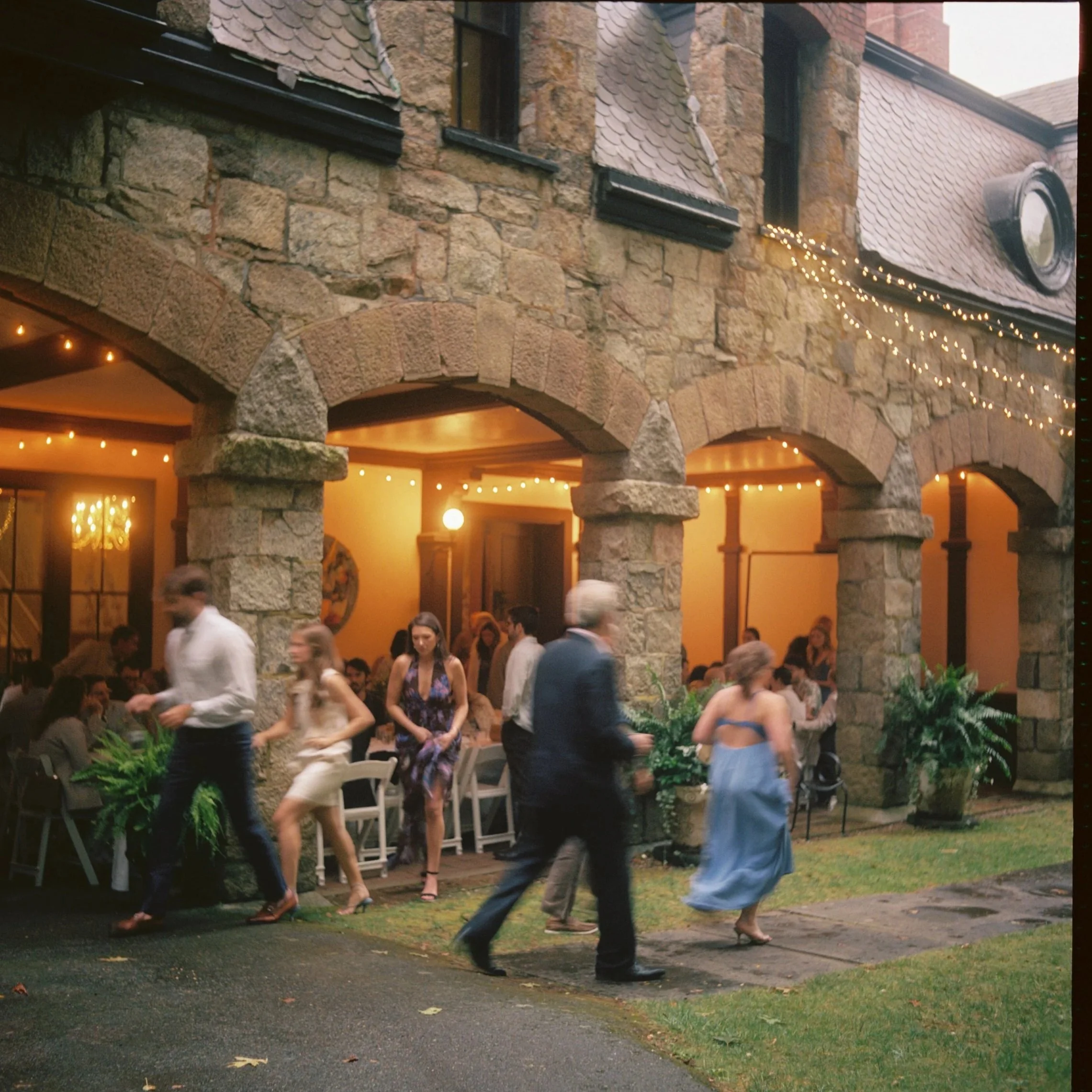 People dressed in formal attire socializing and dancing outside a rustic stone building decorated with string lights during a celebration or wedding reception. Film wedding photographer in Maine. 
