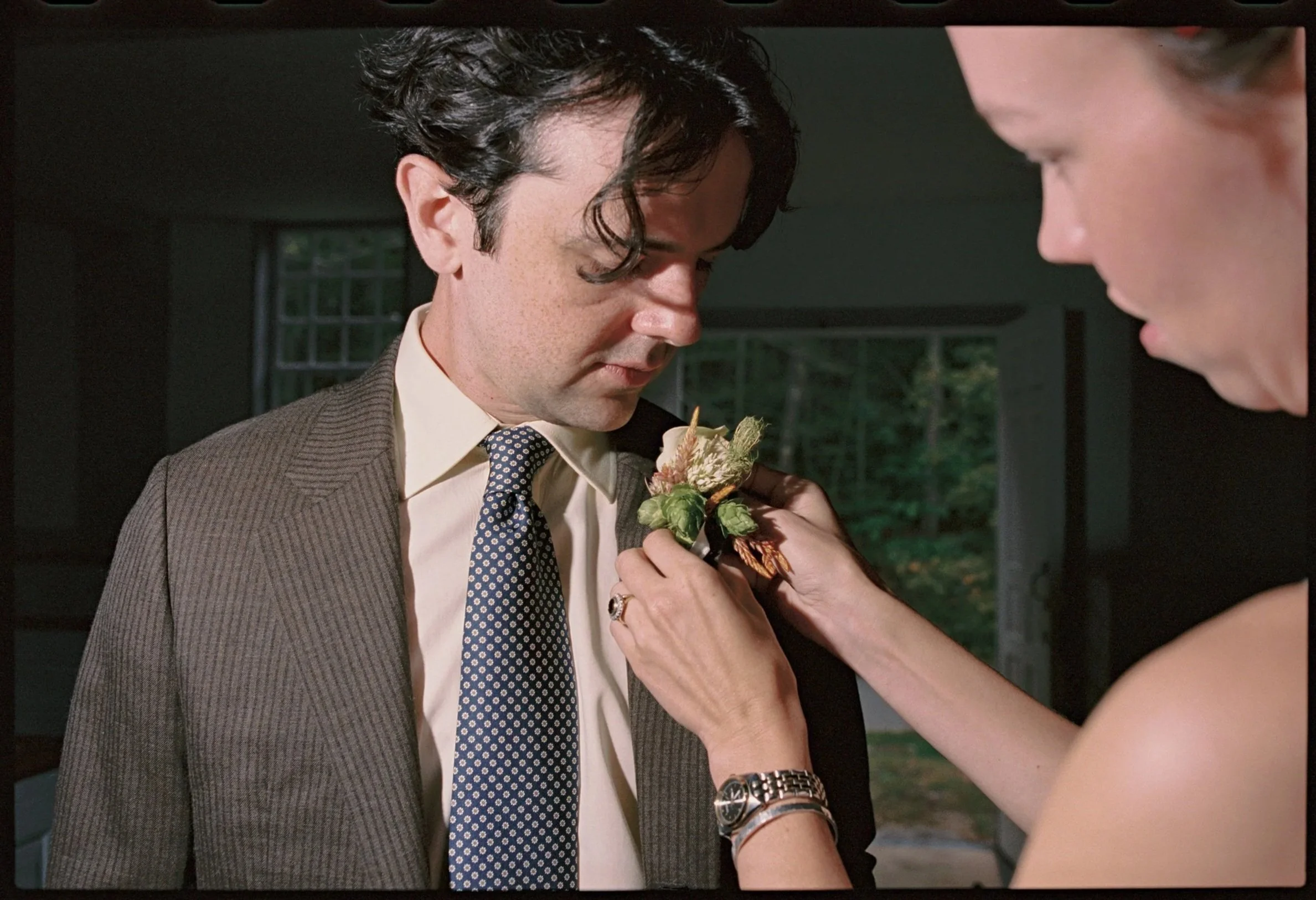 A woman pins a boutonniere onto a man's suit jacket during a wedding or formal event, in an indoor setting with windows and greenery outside. Film wedding photographer in Maine. 