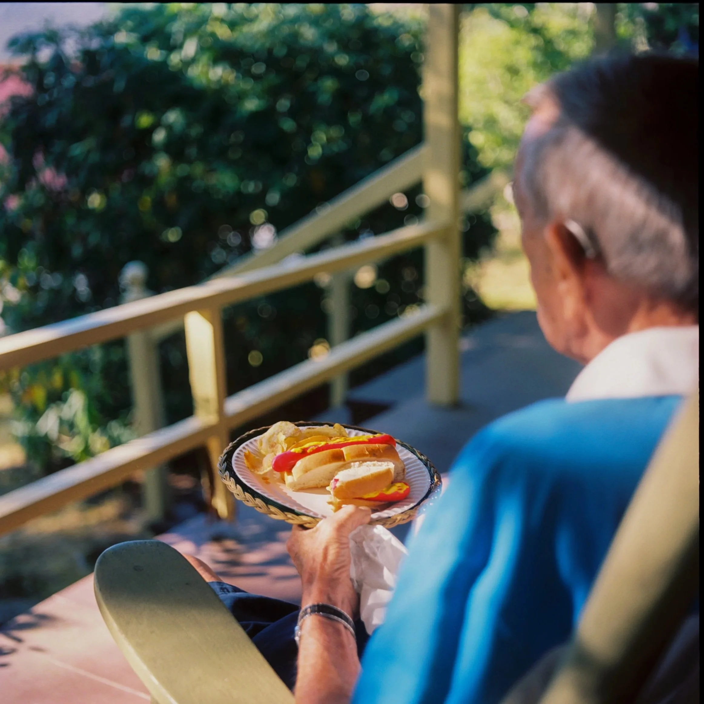 An elderly man sitting outside on a porch, holding a plate with hot dogs and potato chips, with greenery and stairs in the background. Non -traditional film wedding photographer in Maine. 
