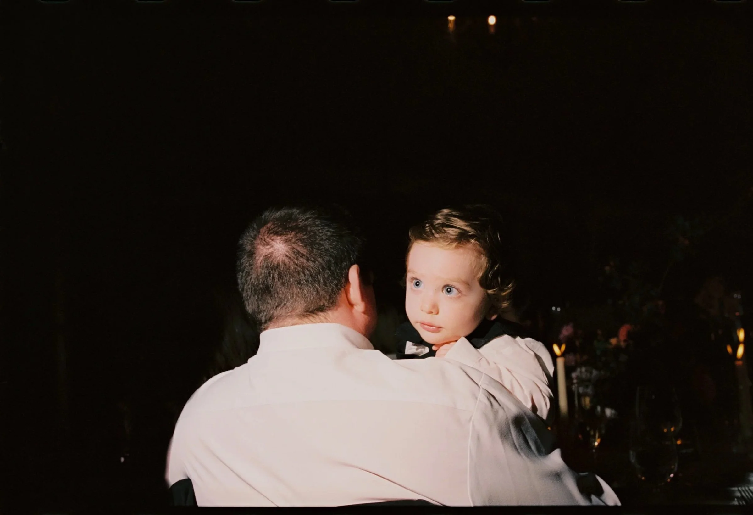 A man holding a young child with curly hair and bright blue eyes, indoors at night with a dark background and blurred lights.