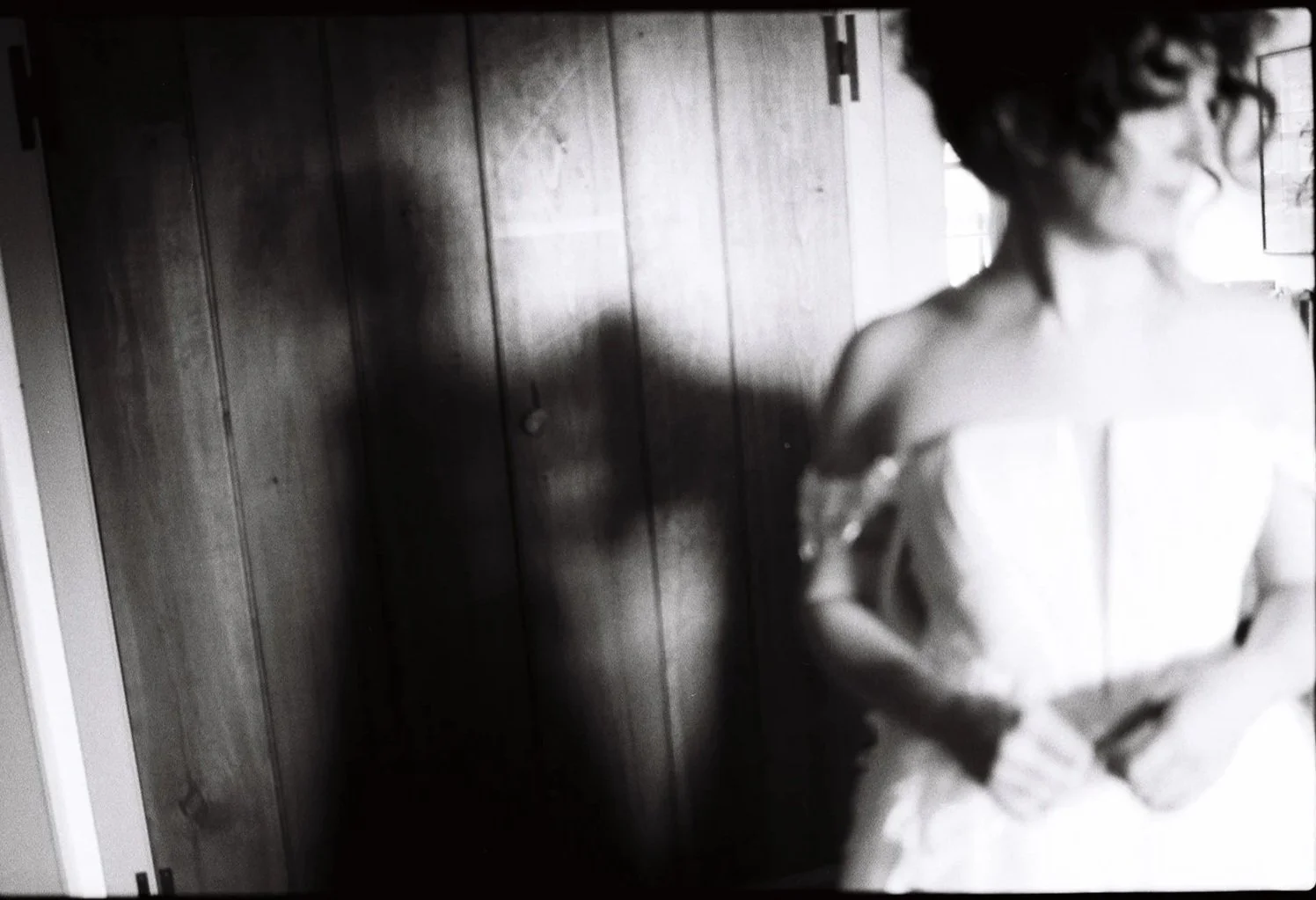 A black and white photo of a woman wearing glasses and an off-the-shoulder dress, standing in front of a wooden wall, with her head turned to the right and her arms crossed.Non -traditional film wedding photographer in Maine. 