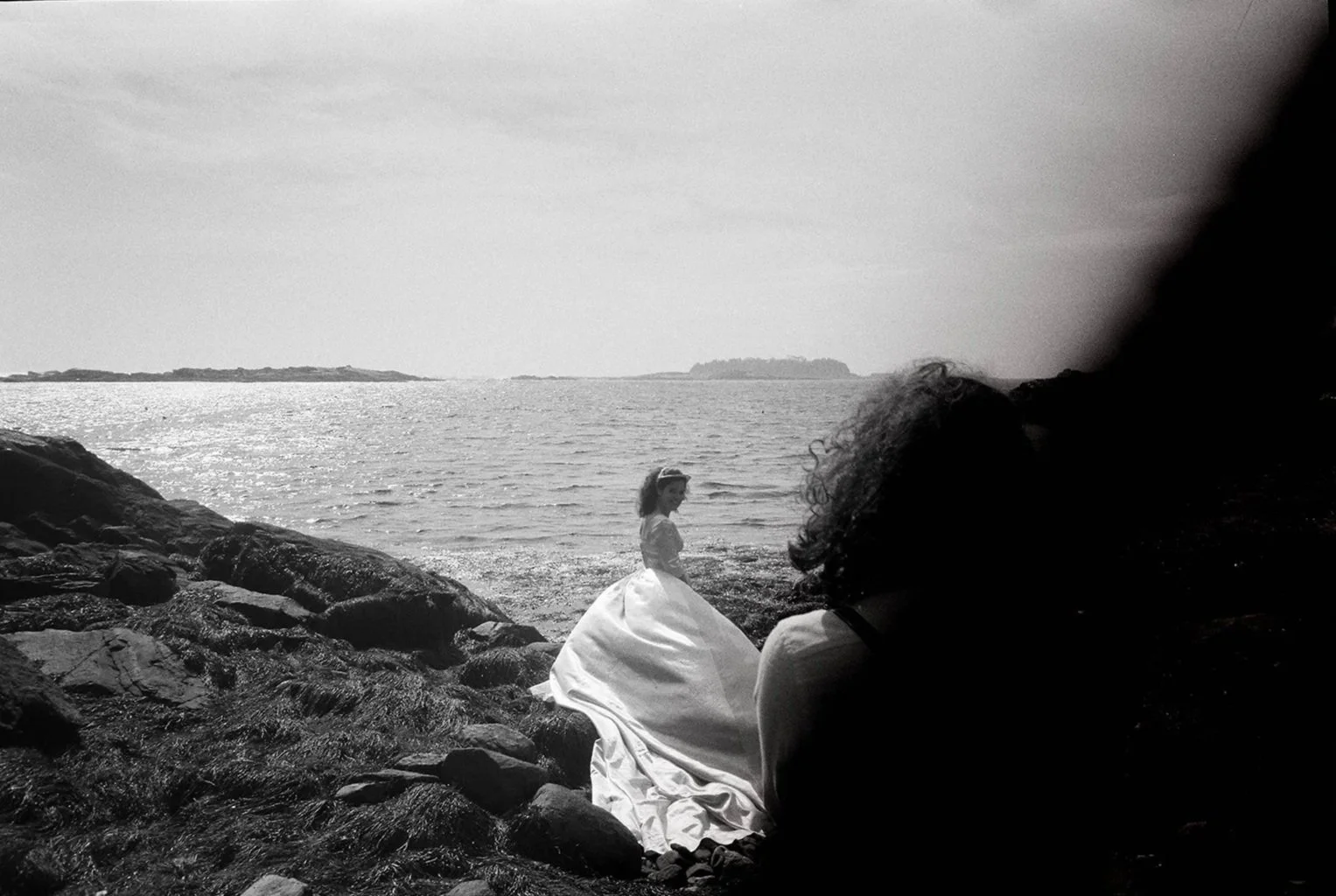 A woman in a white dress standing on rocks near the ocean with another person sitting nearby.