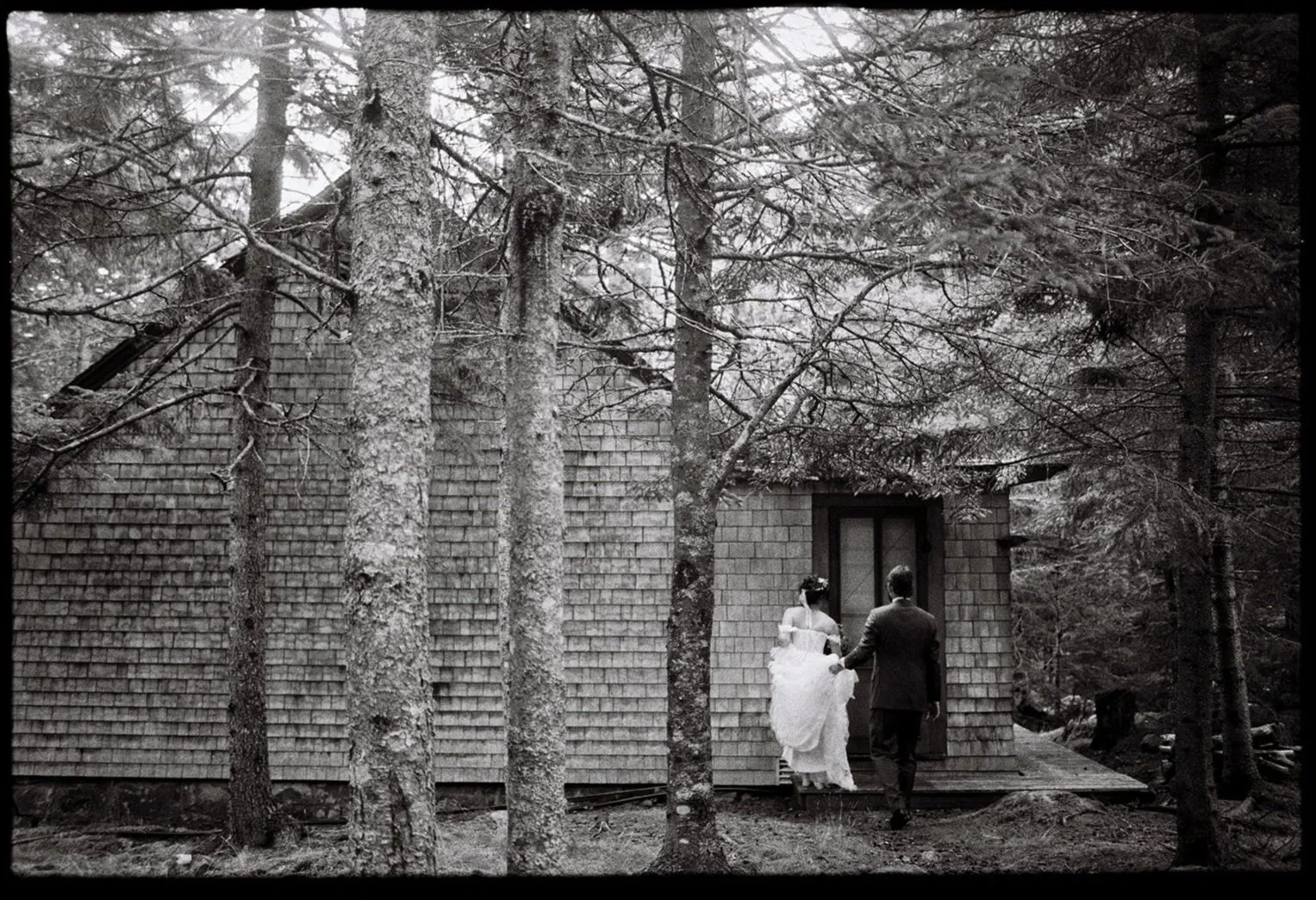 A black and white photo of a couple walking towards a house in a forest, with tall trees surrounding the building. Non -traditional film wedding photographer in Maine. 