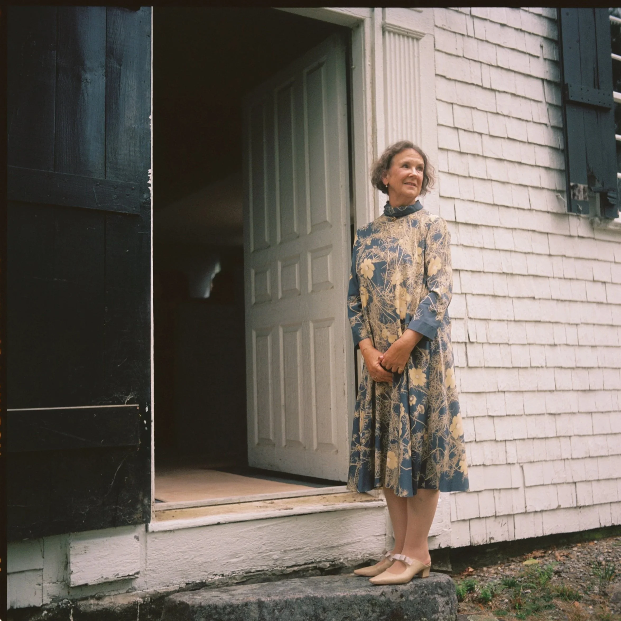 An elderly woman in a floral dress and beige shoes standing on a stone step outside a white wooden house with blue shutters and a open door.