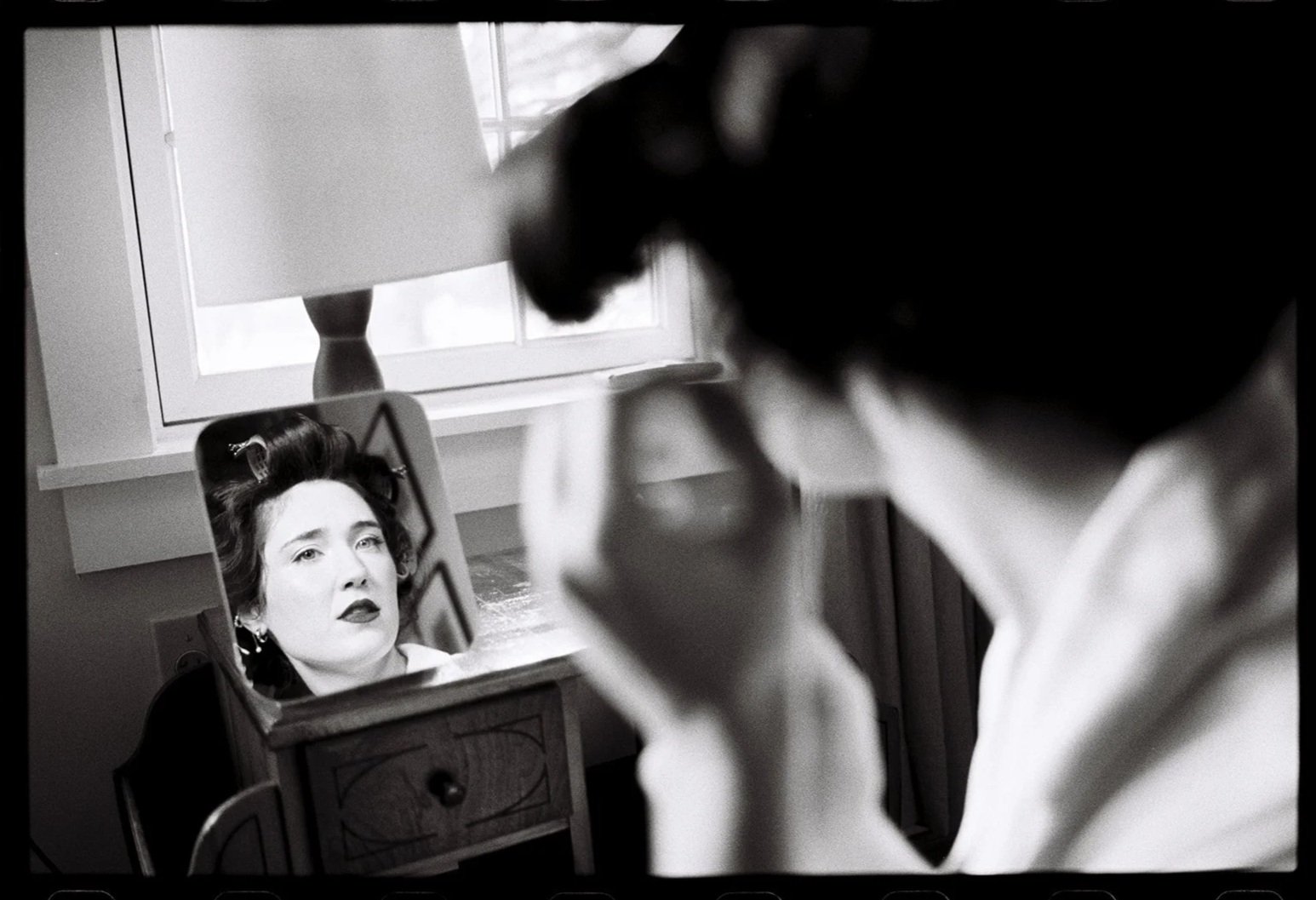 A woman looking at herself in a mirror as a makeup artist applies makeup to her face in a vintage-style setting. Non -traditional film wedding photographer in Maine. 