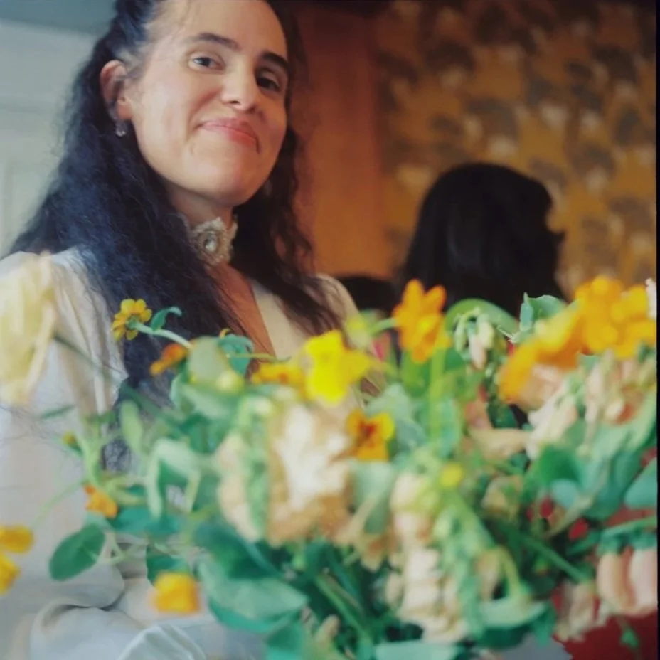 A woman with long dark hair smiling, seated at a table with a floral arrangement of yellow and white flowers in front of her, in a warmly lit indoor setting with a stone wall in the background. Non -traditional film wedding photographer in Maine. 