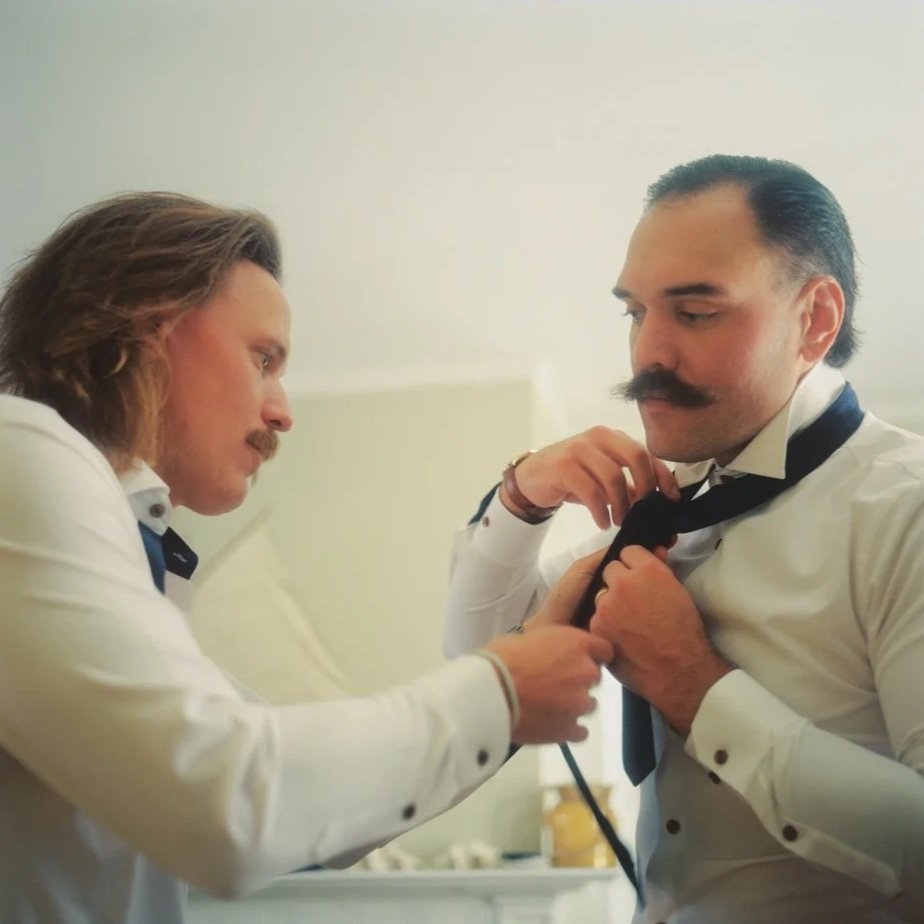 Two men with mustaches wearing white dress shirts and black ties, with one helping the other tie his tie. Non -traditional film wedding photographer in Maine. 