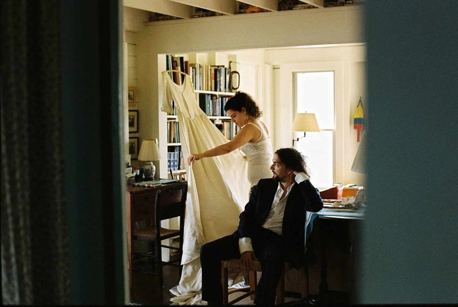 A woman is holding a dress, standing on a chair, next to a seated man inside a cozy room with bookshelves and large windows.