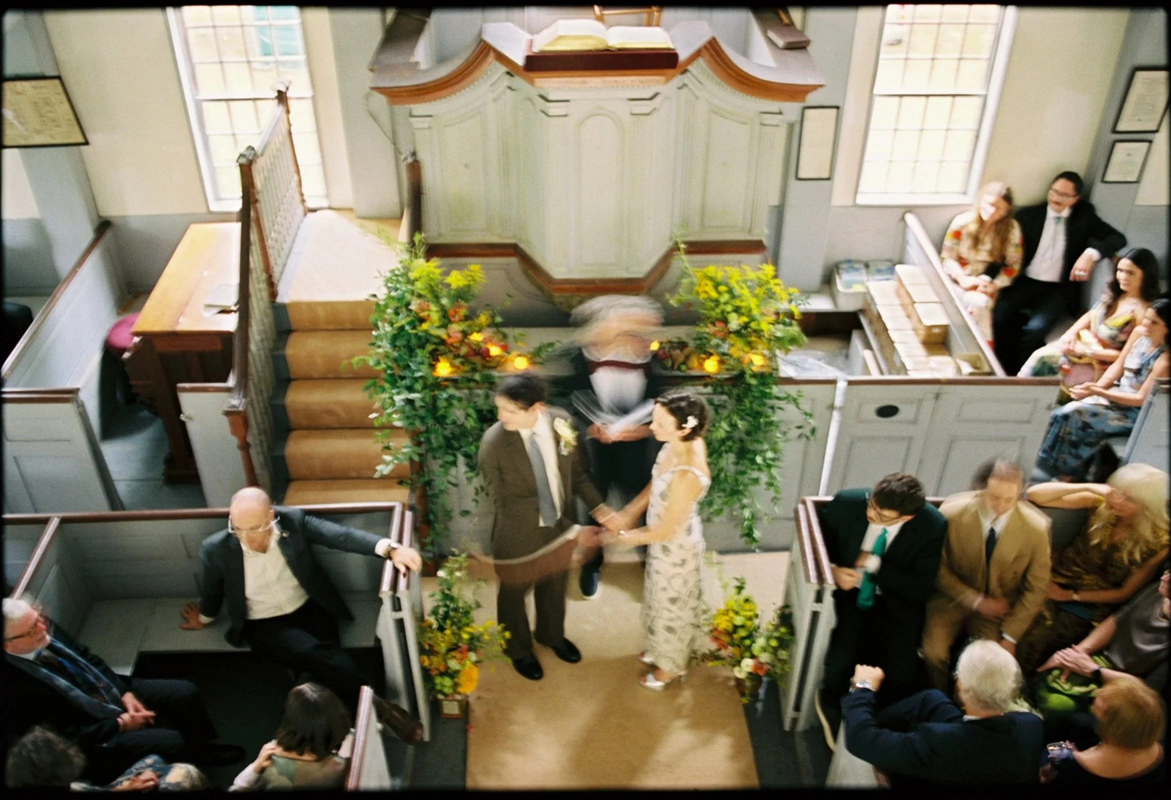 A wedding ceremony in a church, with a bride and groom holding hands in the center, surrounded by guests seated in pews and standing, in a well-lit room with large windows and religious decorations.