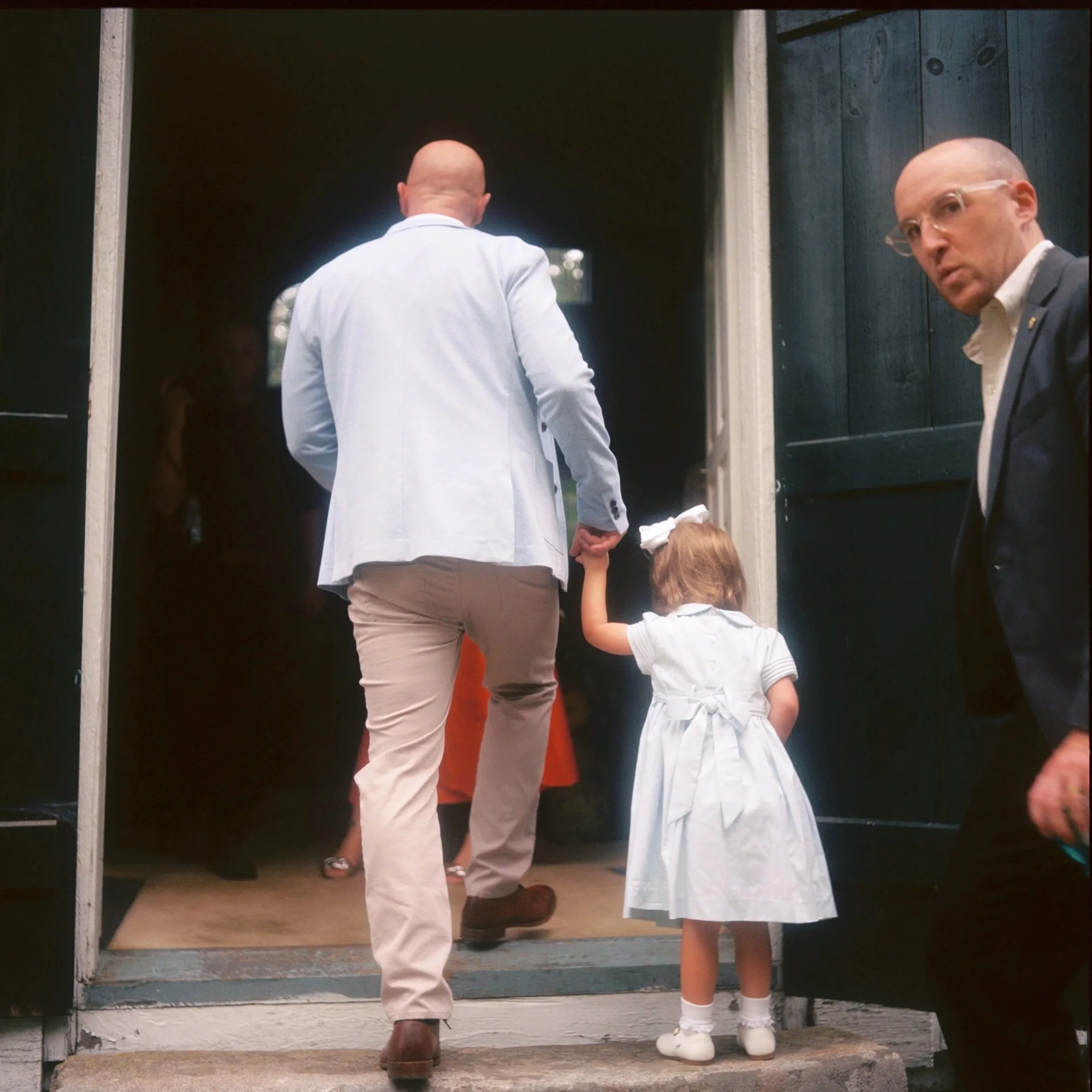 A man in a light blue jacket holding a girl's hand as they walk into a building, with another man in a dark suit observing from the side. Non -traditional film wedding photographer in Maine. 