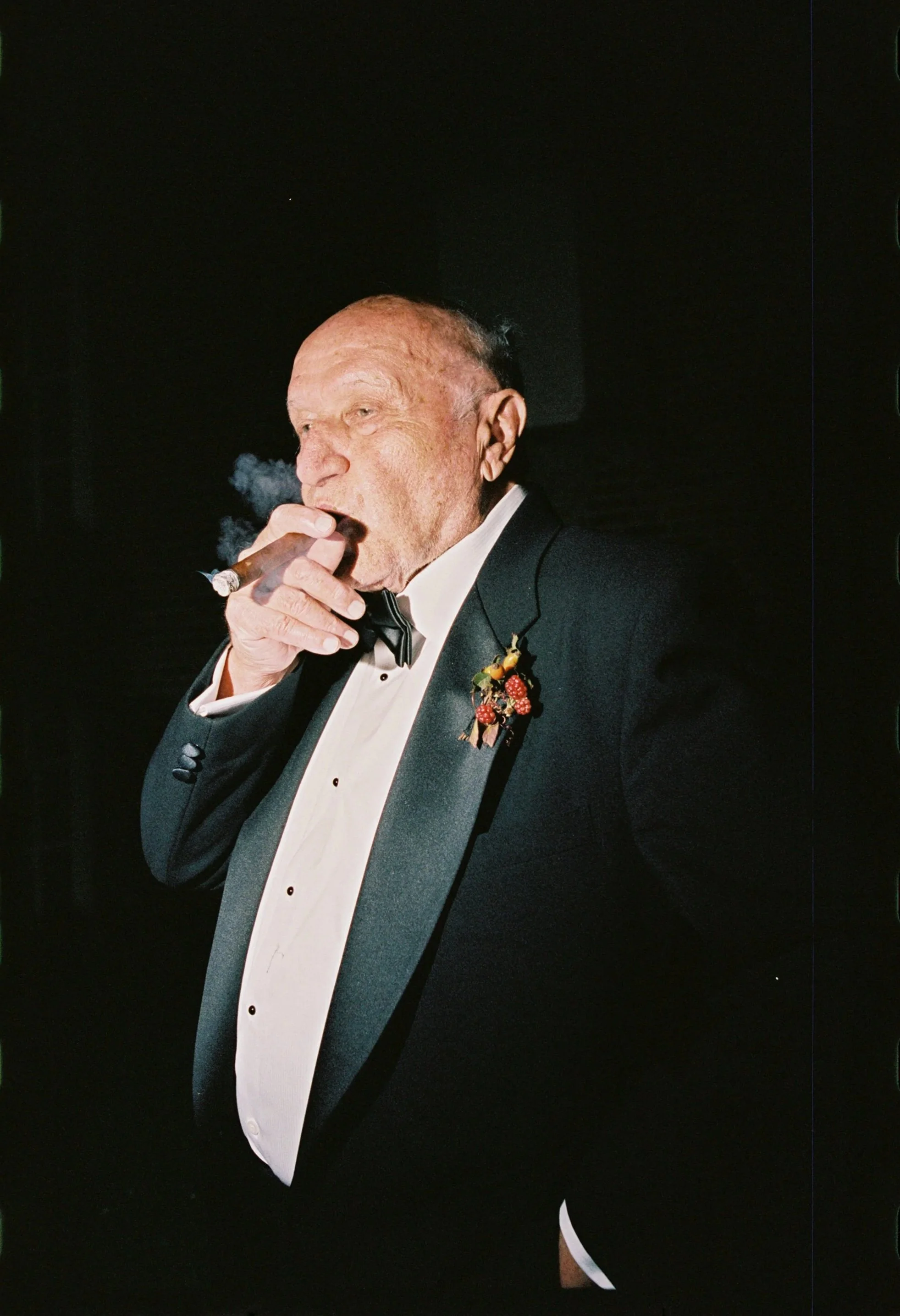 An elderly man in a tuxedo with a bowtie, smoking a cigar at a formal event, with a boutonnière on his lapel. Non -traditional film wedding photographer in Maine. 