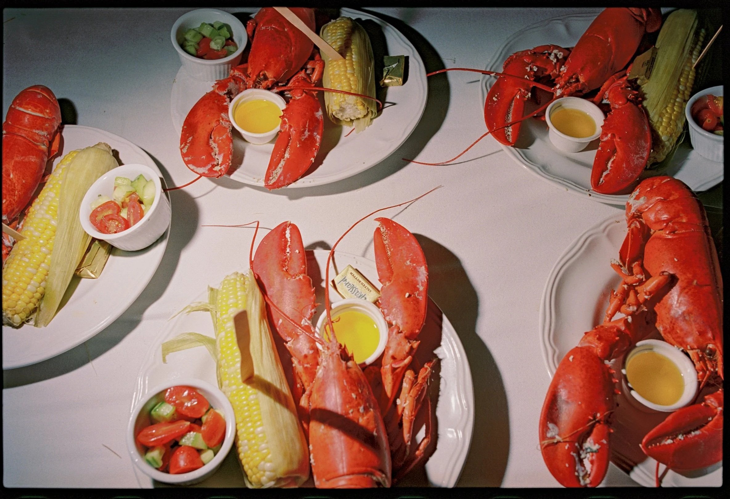 Lobster boils served on white plates with corn on the cob, small bowls of melted butter, and salads with cherry tomatoes and cucumbers. Non -traditional film wedding photographer in Maine. 